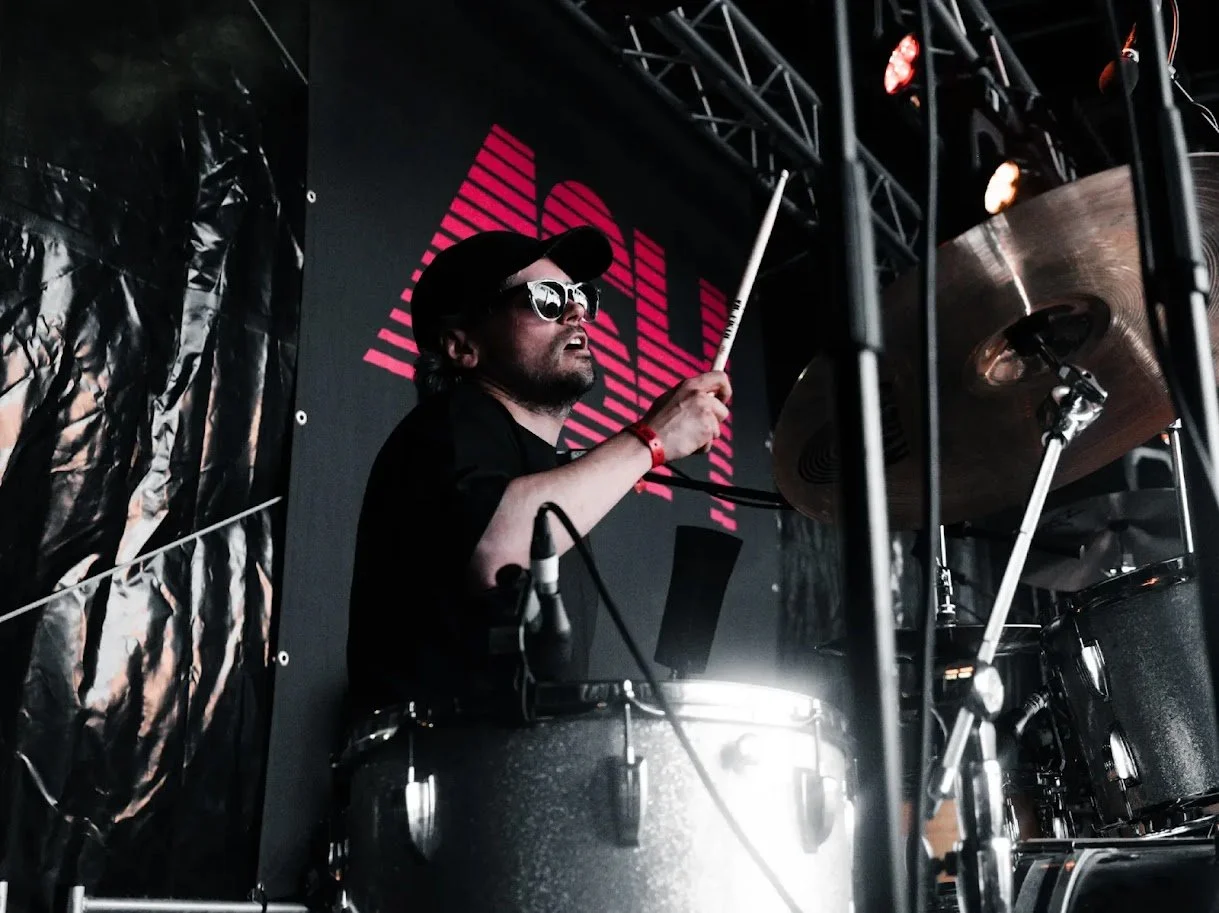 A male drummer wearing sunglasses, a black baseball cap, and a black shirt playing drums on stage with a dark background and a large red and pink graphic behind him.