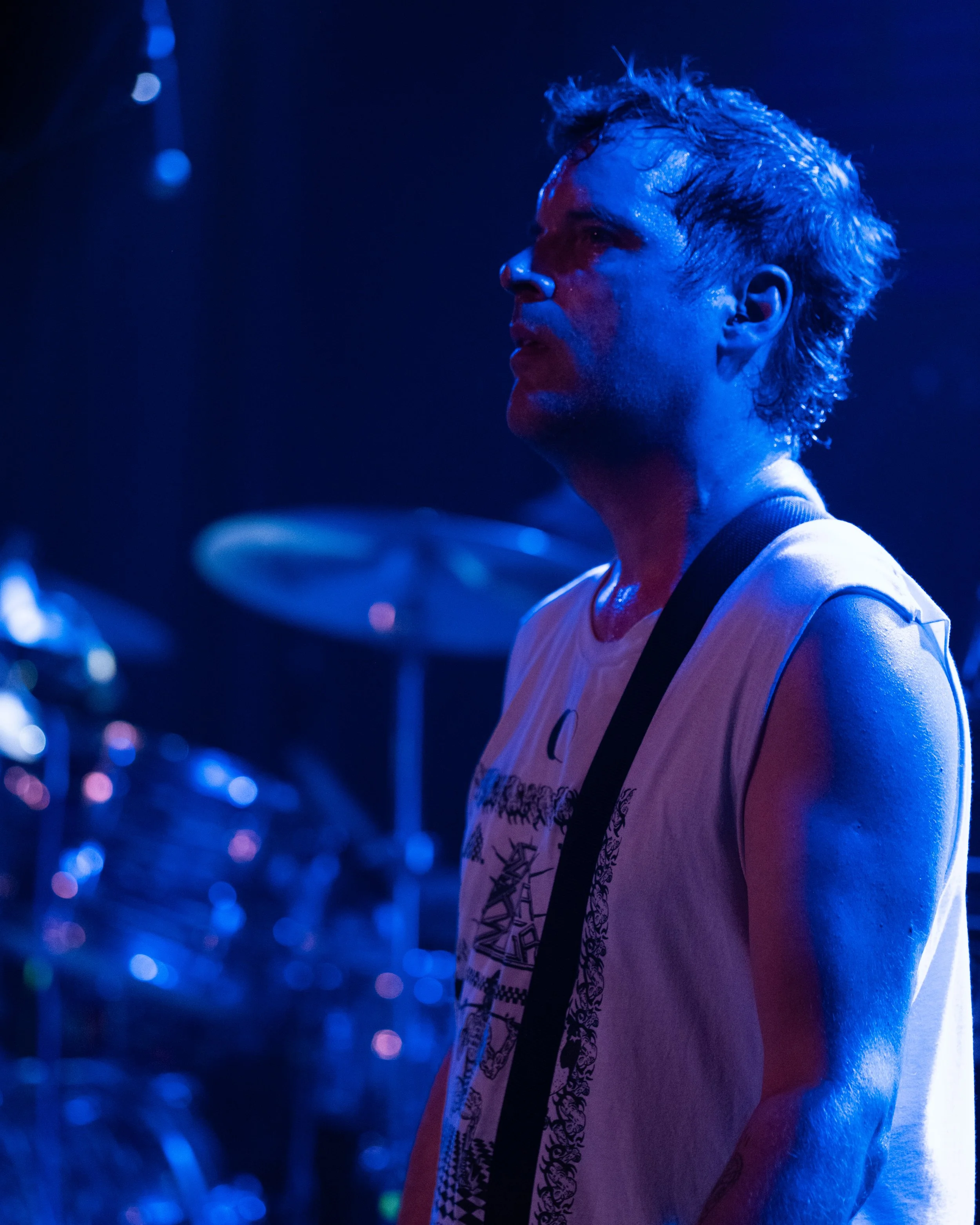 A male musician standing on stage with wet hair, illuminated by blue stage lighting, with drums in the background.