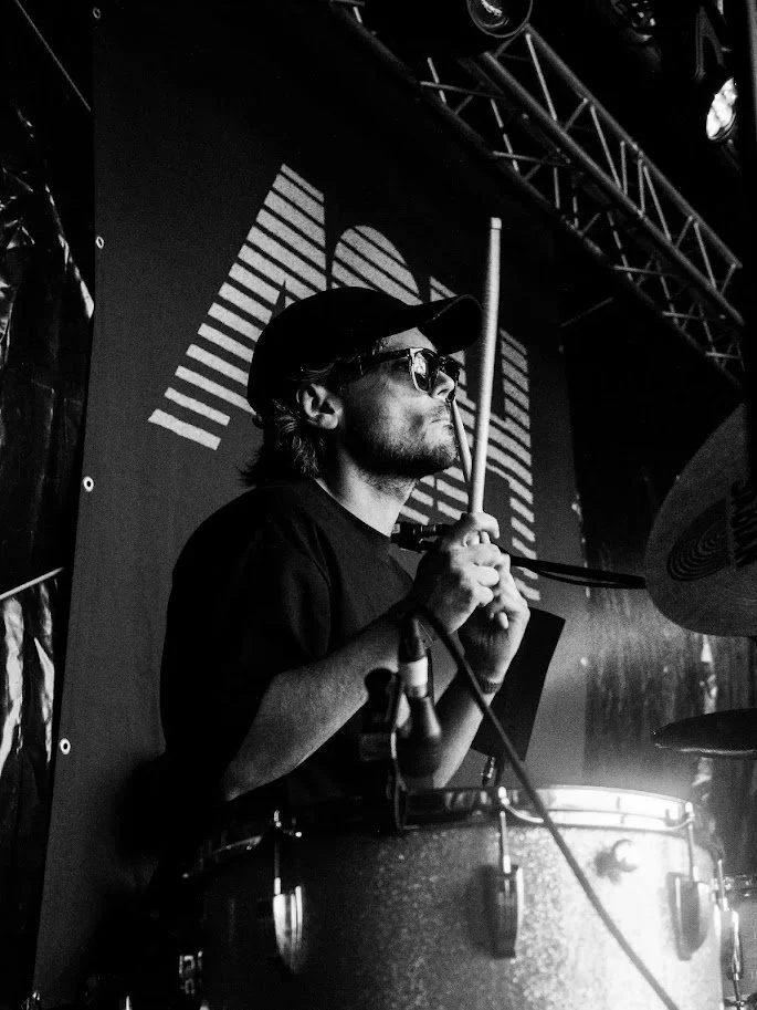 A man wearing sunglasses and a cap is playing drums on stage, with a large screen behind him displaying a geometric pattern.
