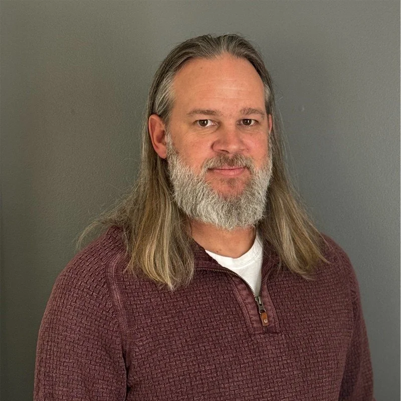 A smiling man with a beard wearing a gray beanie and a dark jacket, sitting indoors with other people in the background.