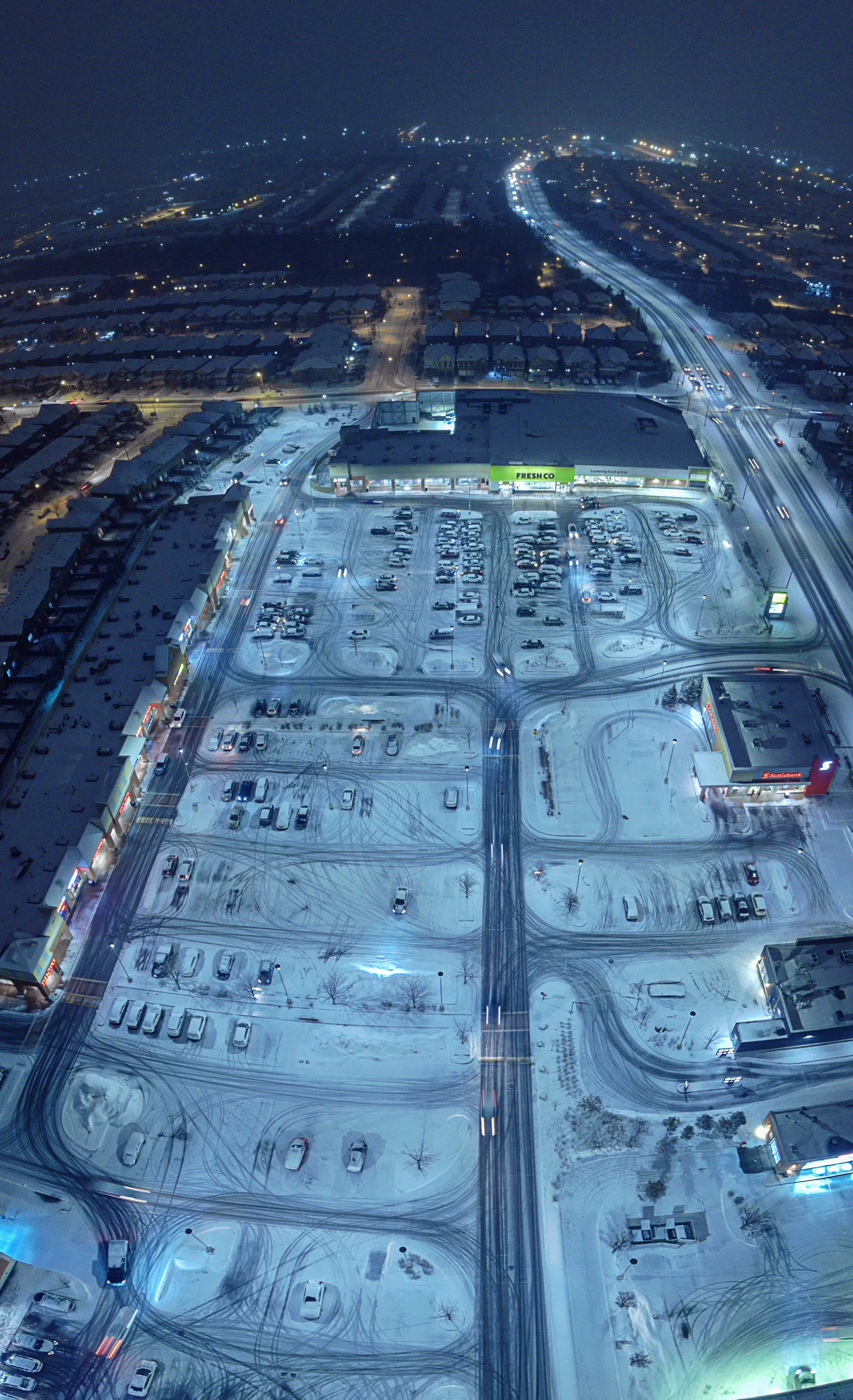 "Drone-captured vertical panorama during a snowstorm, showcasing the intricate paths etched by vehicles in the snow—tracing motion and texture across a wintry urban canvas."