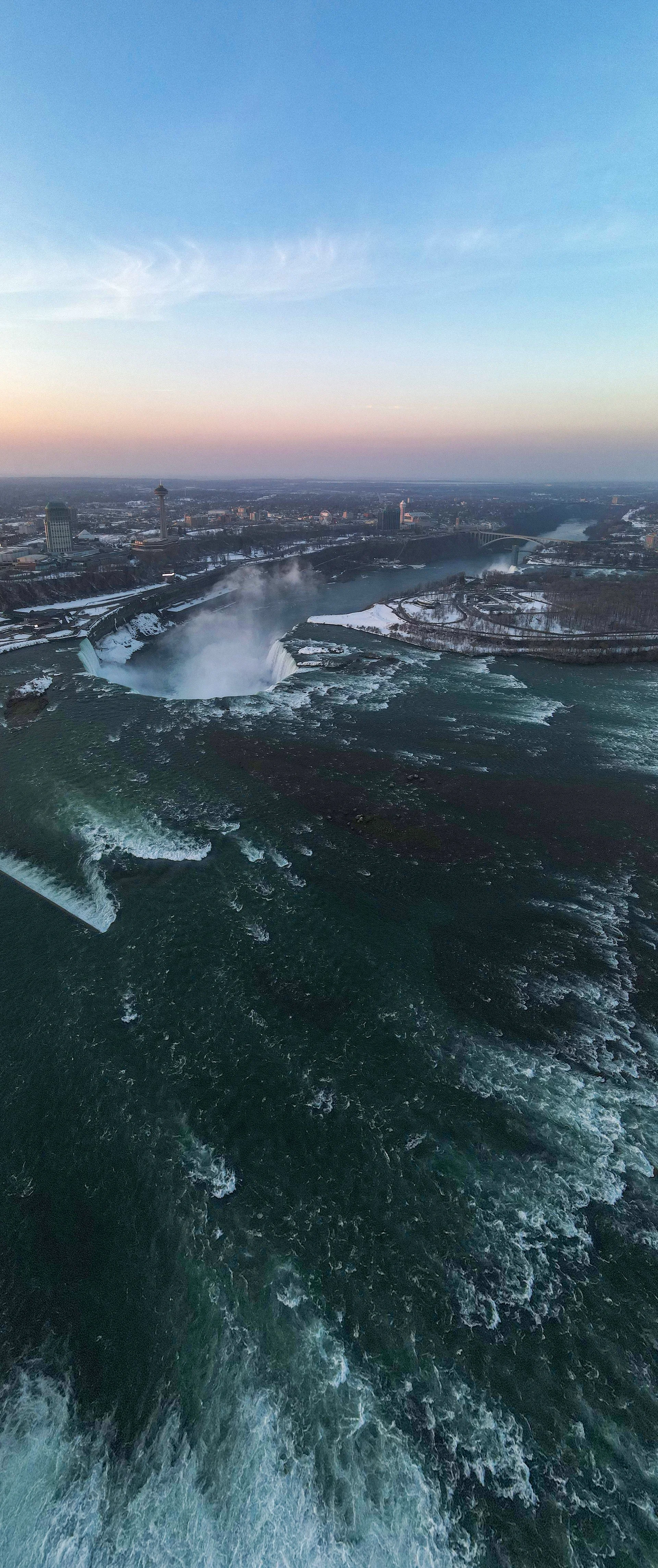 "Vertical panoramic view of Niagara Falls on the Canadian border, captured from a drone to reveal the dramatic drop and mist rising from the cascading water."