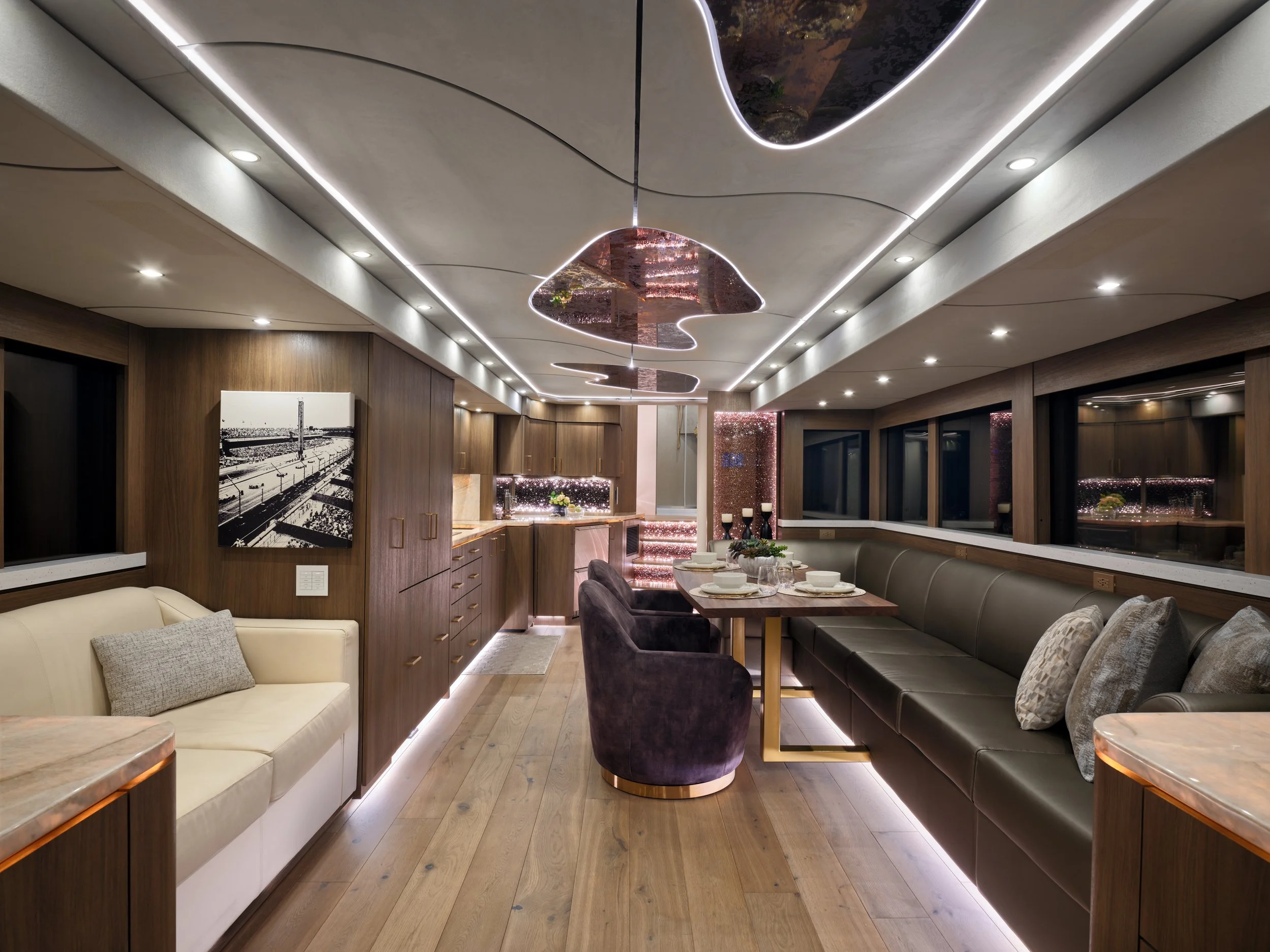 Modern dining room with wooden walls, a long dark leather banquette, purple velvet chairs, and a table set with white dishes and glasses. recessed lighting and decorative ceiling fixtures enhance the space, with large windows along one wall.