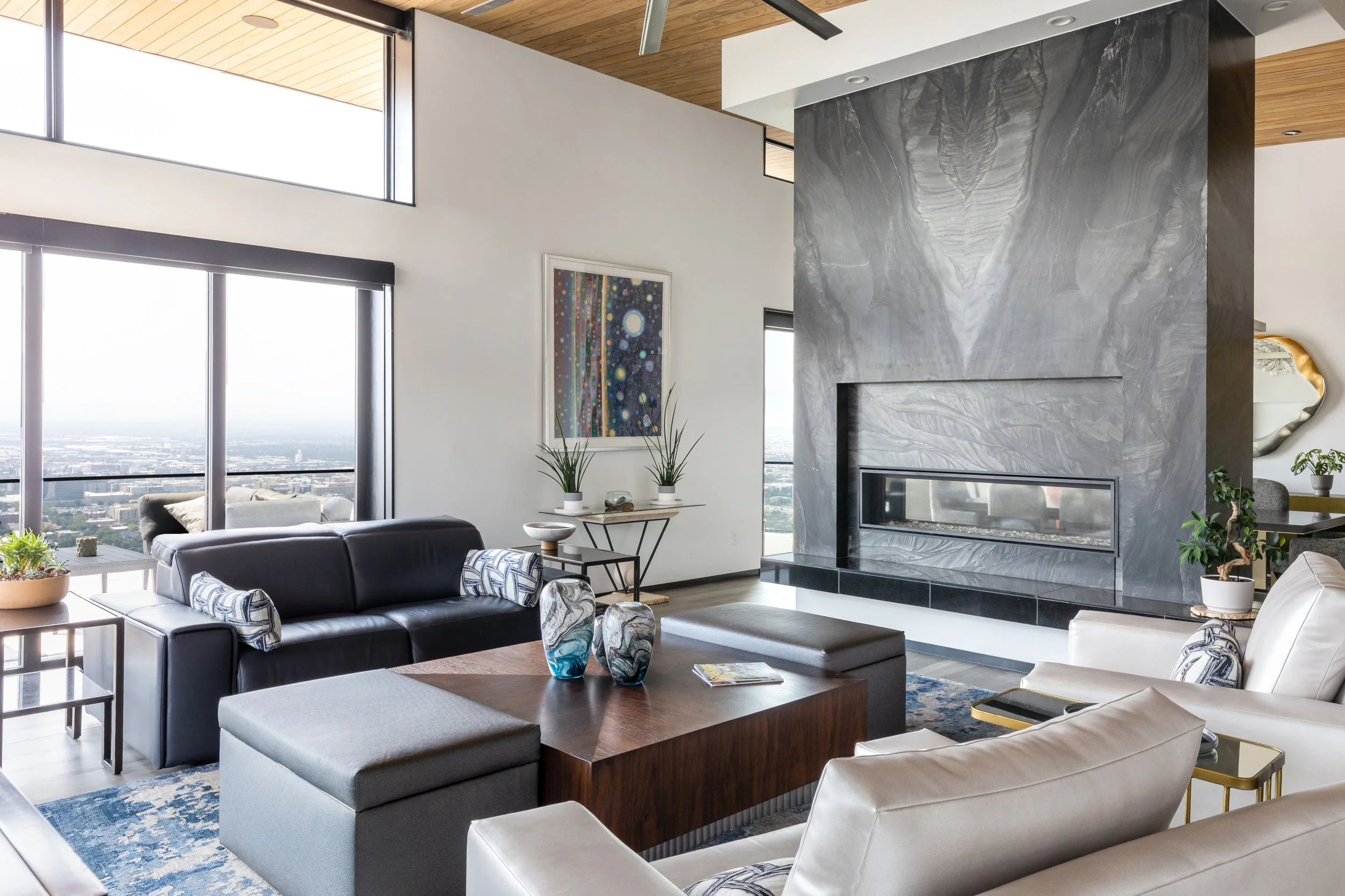 Modern living room with a black leather sofa, white armchairs, a wooden coffee table, a gray marble fireplace, large windows showing a city view, and contemporary art and decor.