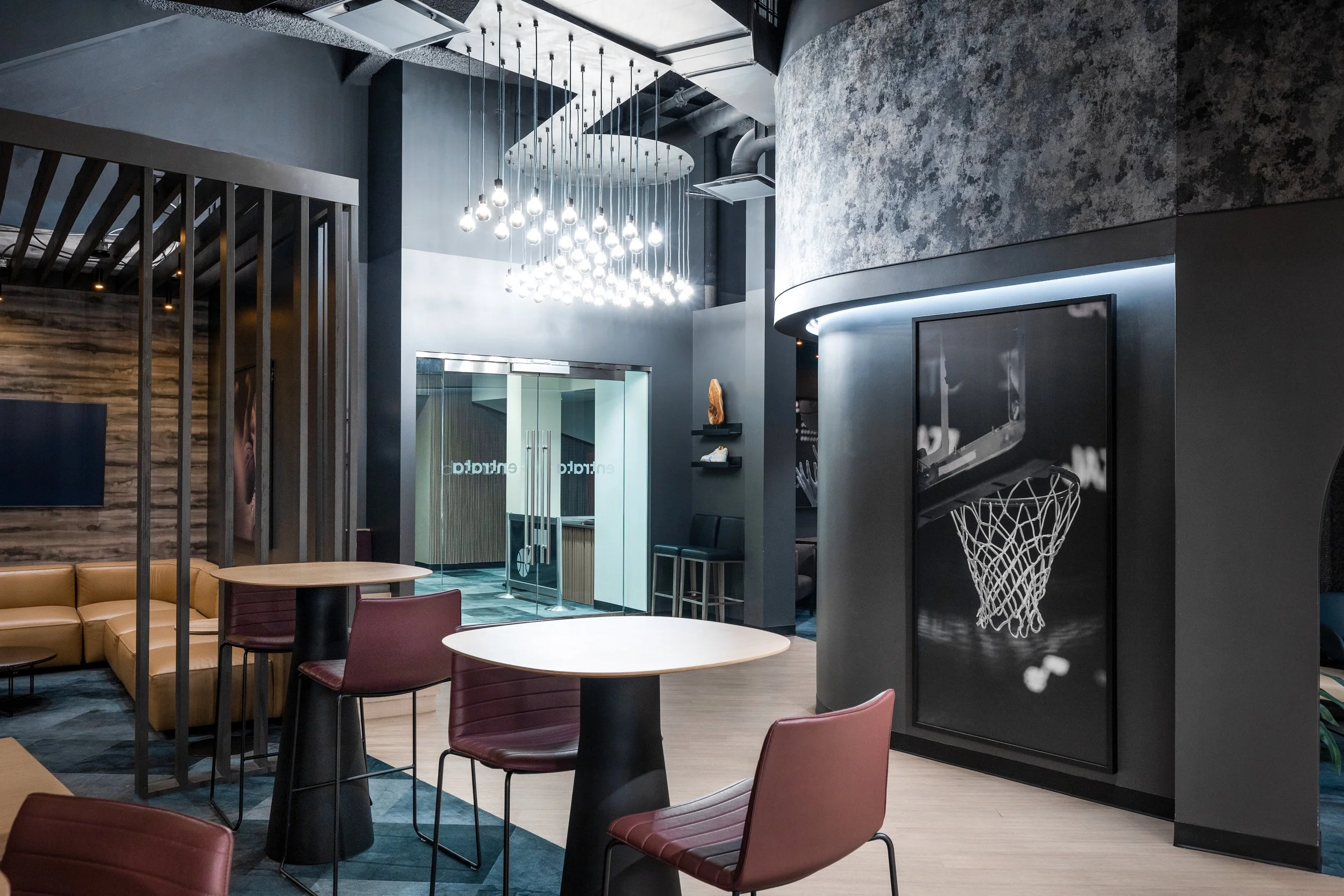 Modern lounge with tables, chairs, a wall-mounted television, and pictures of a basketball hoop and court, illuminated by a contemporary hanging light fixture.