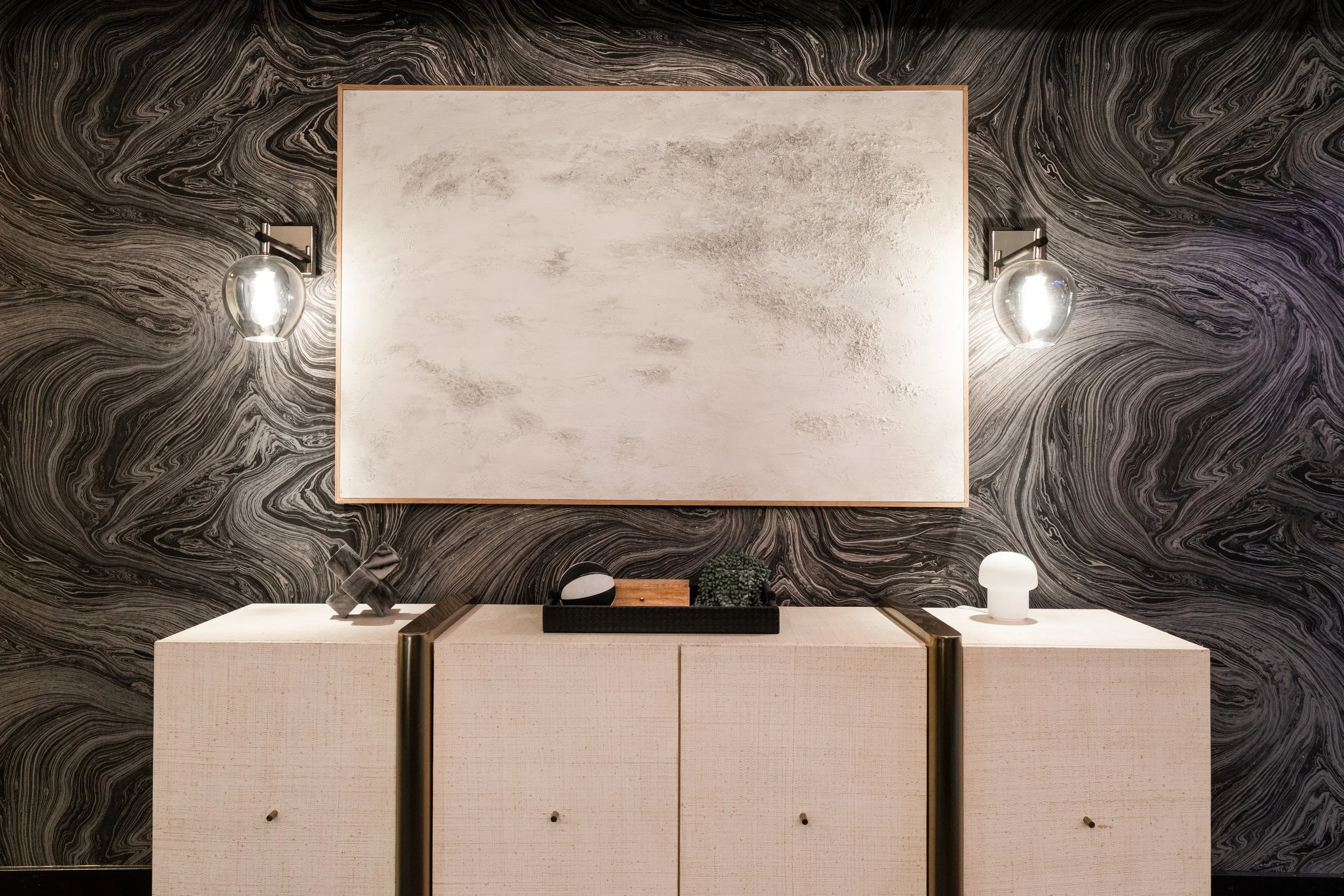 Modern beige sideboard against a black and white marble patterned wall with a large blank white canvas and two glass wall-mounted lamps.
