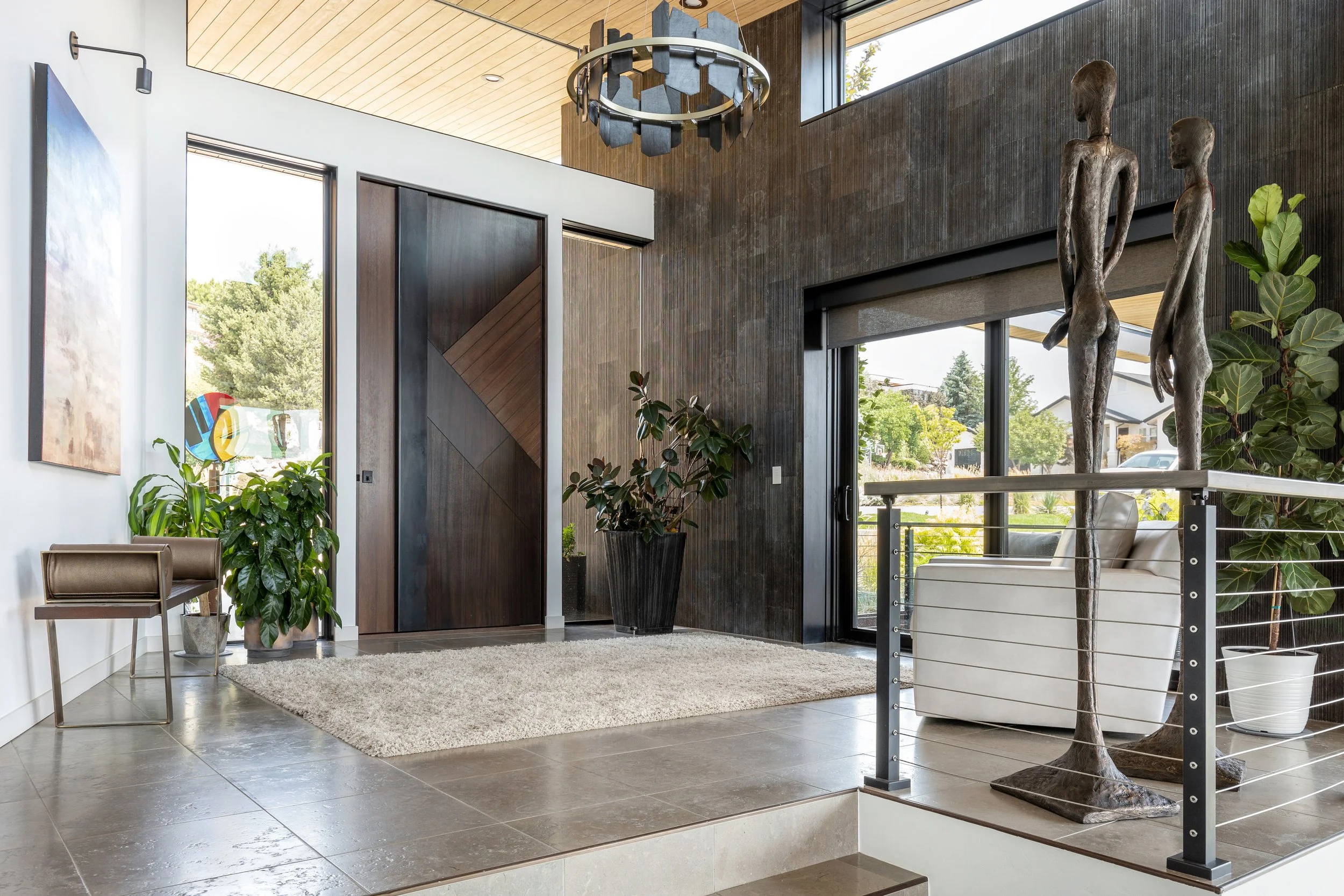 Modern interior entrance area with large glass windows, wooden and dark textured wall, a beige area rug, two tall bronze sculptures of human figures, potted green plants, a cream-colored armchair, and a decorative chandelier.