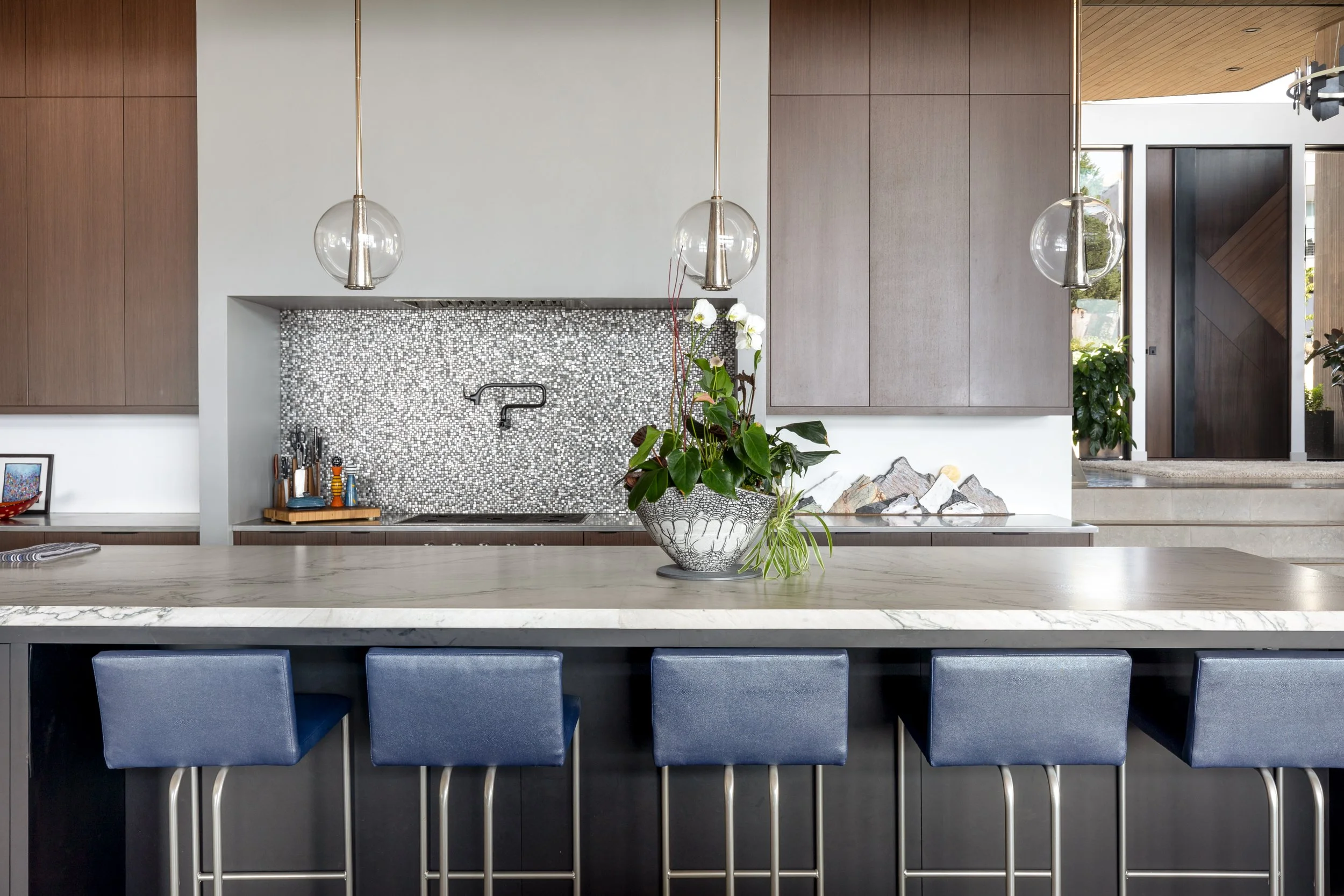 Modern kitchen with a large island, four blue bar stools, a potted plant in the center, and minimalist cabinetry and backsplash.