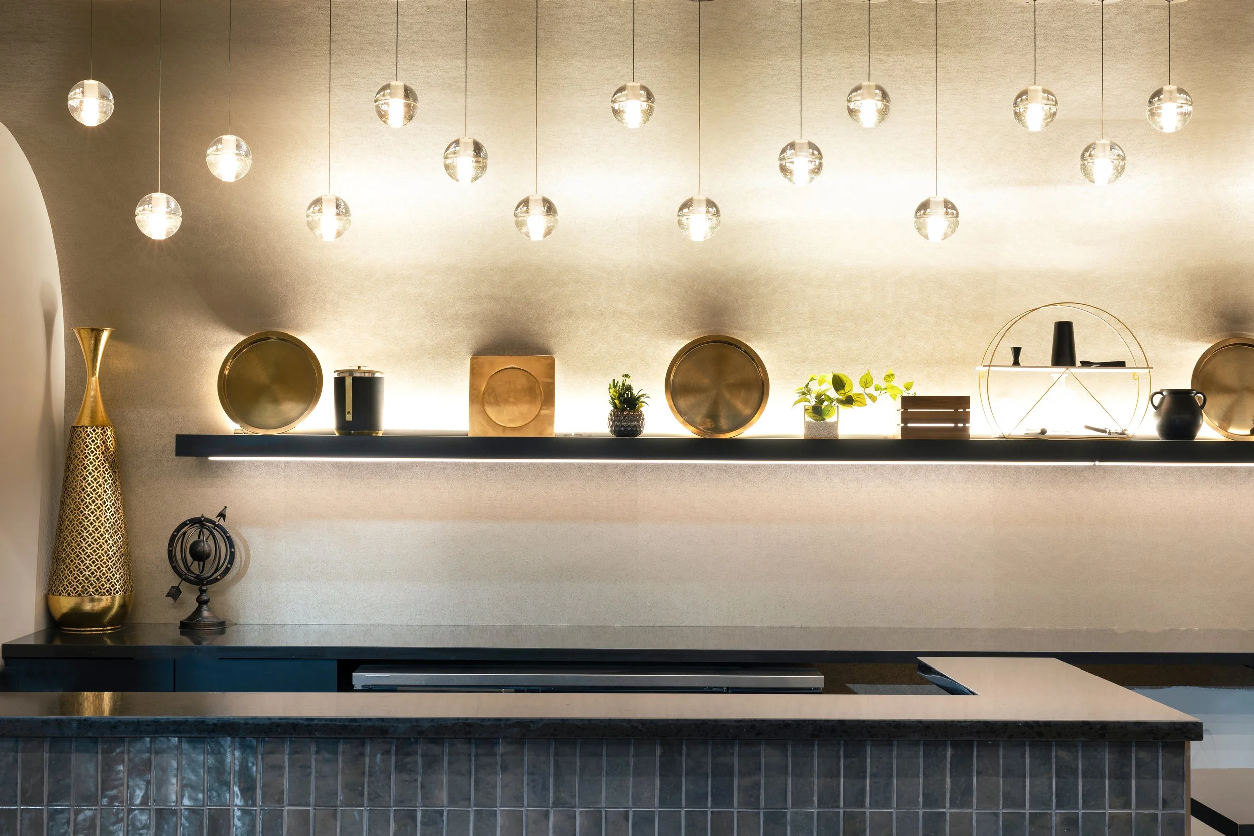 Modern kitchen or bar area with a black countertop, decorative objects, and a shelf with small plants and plates illuminated by hanging globe lights.