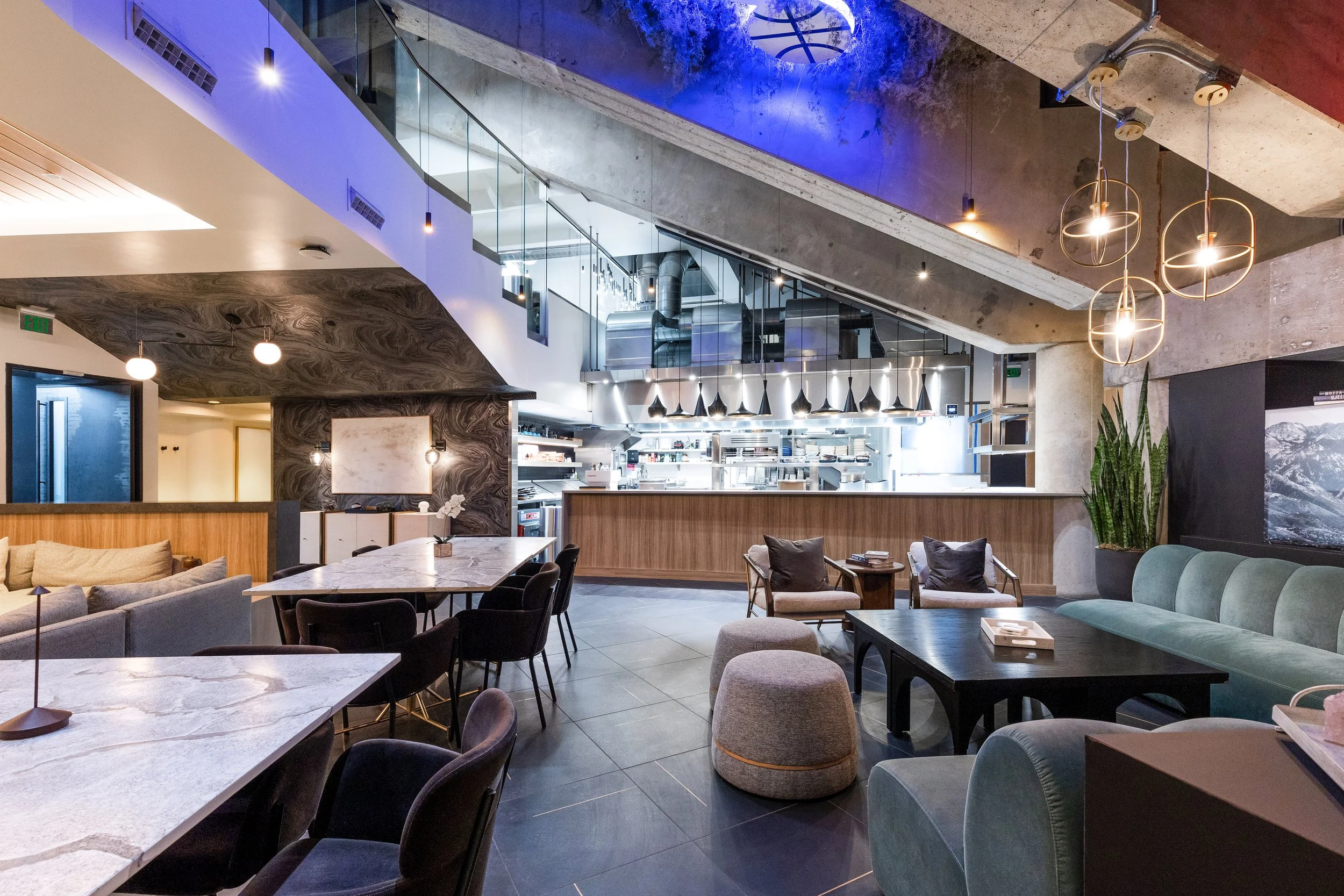 Modern restaurant interior with seating area, open kitchen in background, contemporary lighting fixtures, and decorative plants.