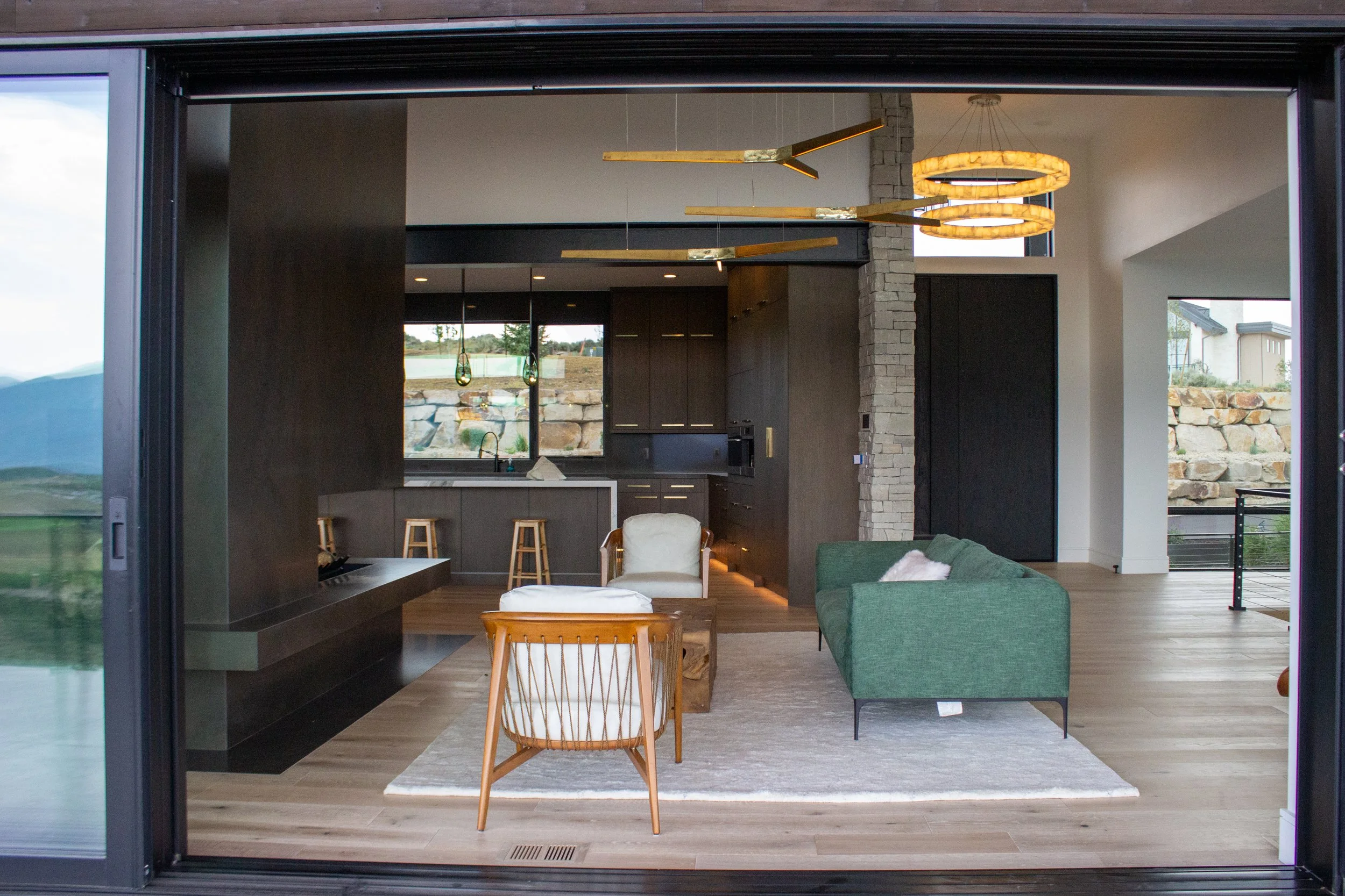 Open living room with a green sofa, white armchair, wooden chair, area rug, kitchen with bar stools, large windows, and modern lighting fixtures.