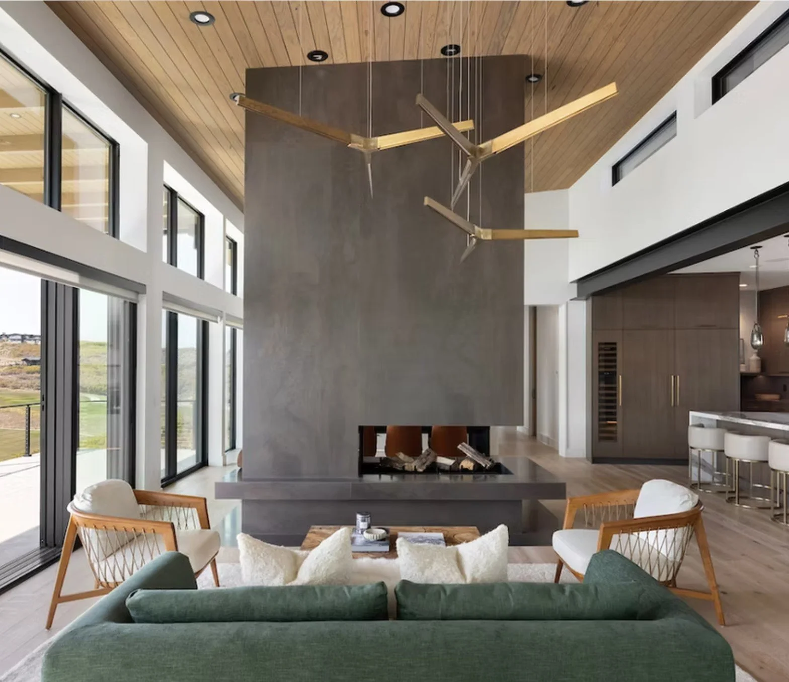 Modern living room with large windows, green sofa, armchairs, and a fireplace with a concrete chimney.