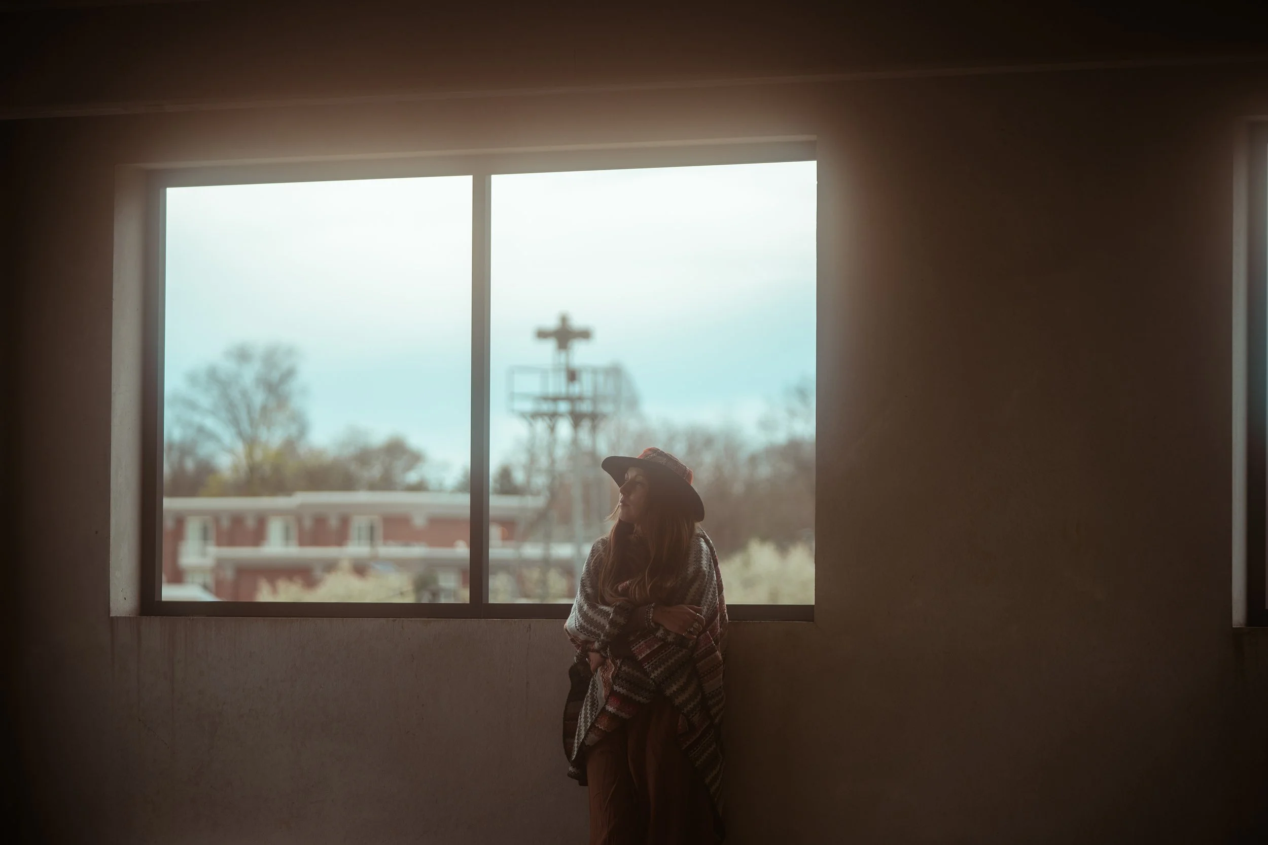 Louka standing in front of a window in a dimly-lit room looking at the ceiling. There's a red building and large structure through the window in the distance.
