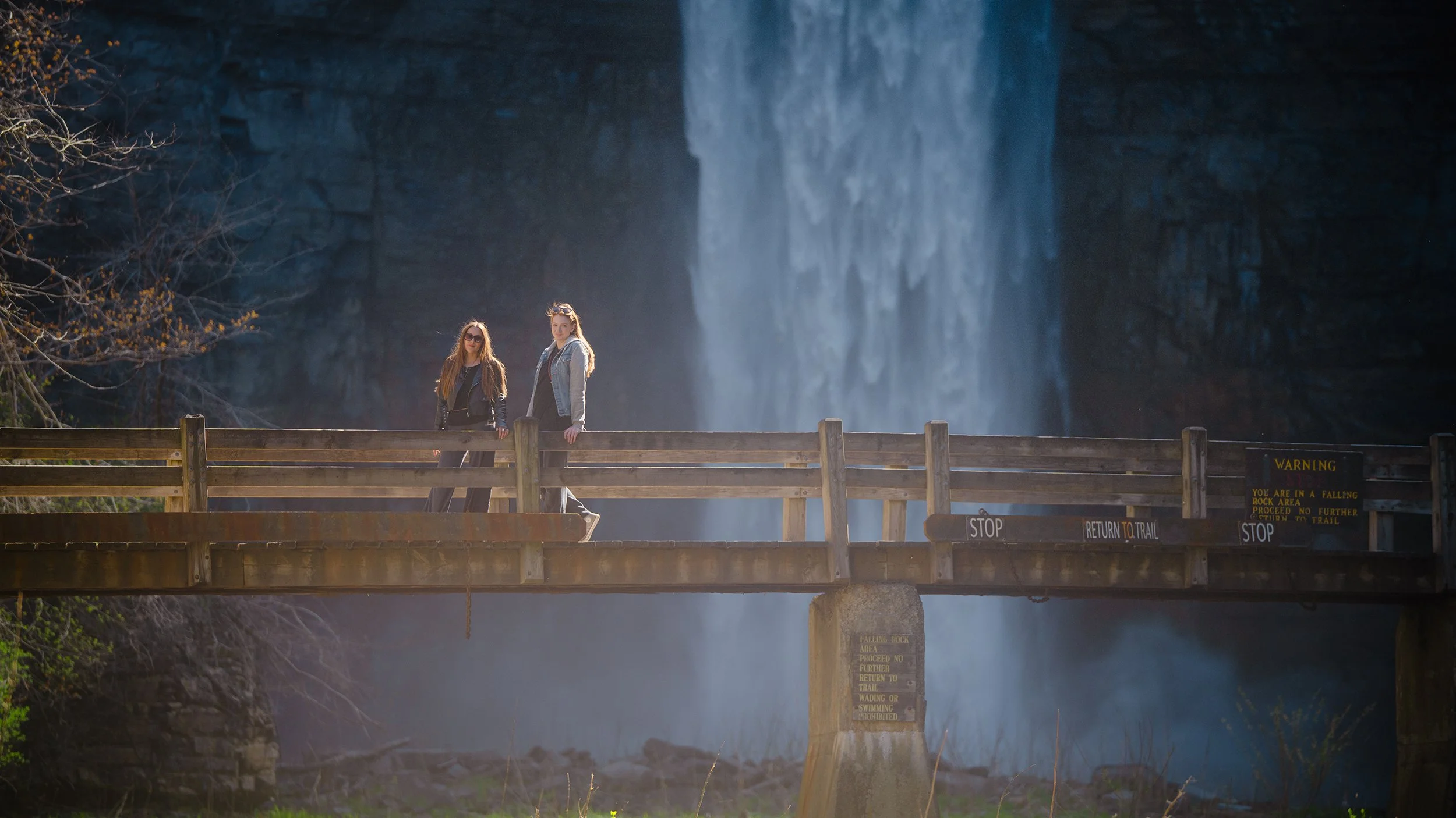 Louka and another woman standing on a suspended wooden bridge with a waterfall in the background.