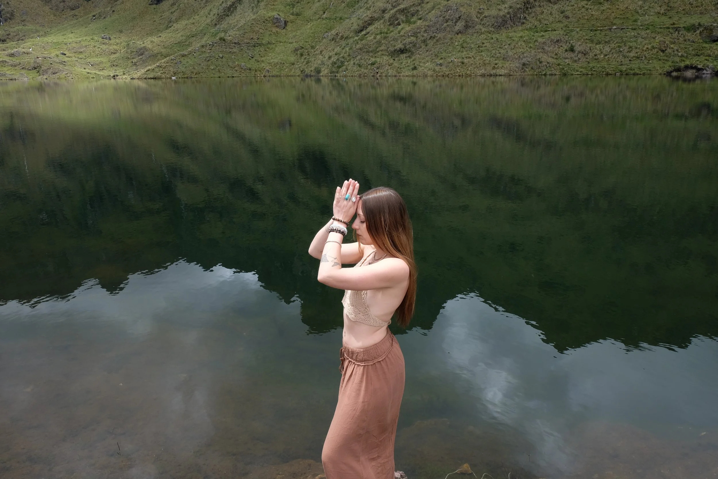 Louka standing next to a body of water in the mountains with her hands held together.