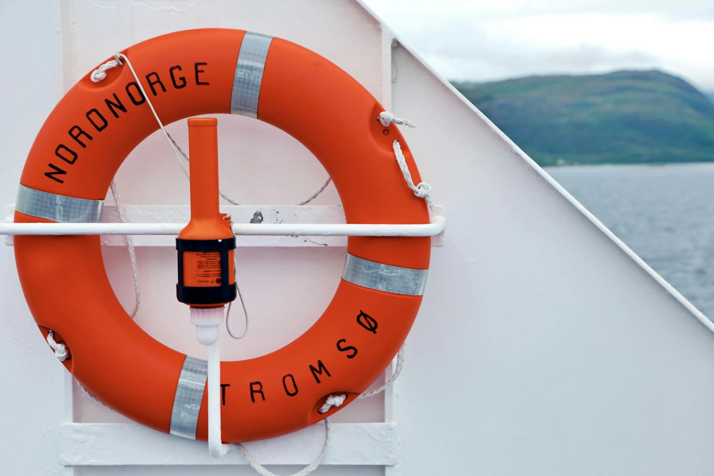 A bright orange lifebuoy with reflective silver stripes and the words 'NOORDNAG' and 'TROMSO' written in black, hanging on the side of a boat, with a Nansen spray nozzle attached. In the background, there's a body of water and a mountainous landscape under a cloudy sky.