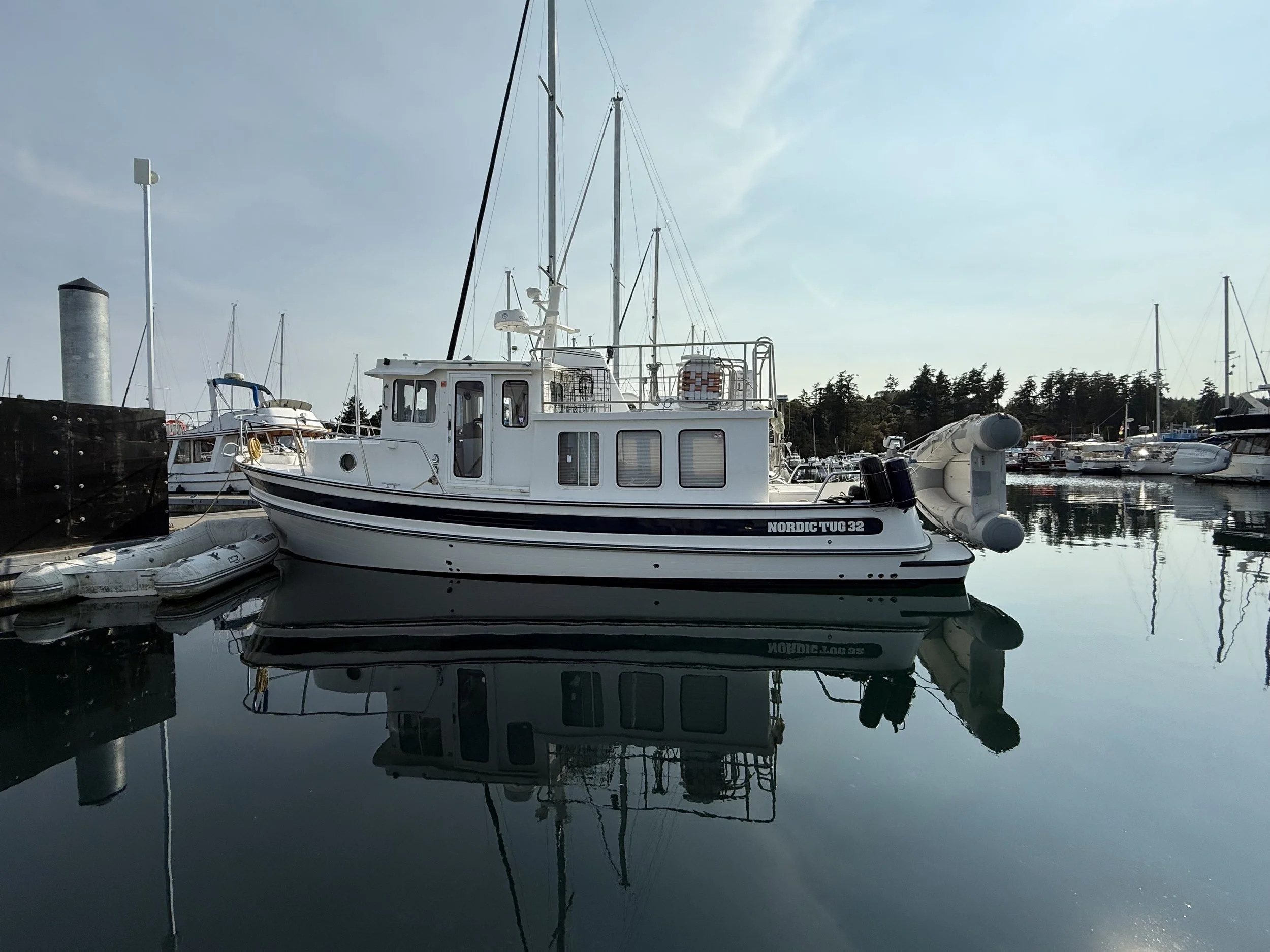 A white boat named Nordic Tug 32 docked at a marina, with other boats and sailboats in the background, and their reflections visible in the calm water.