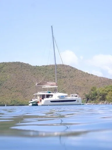 A sailboat with a large mast floating on calm water with hills and a partly cloudy sky in the background.