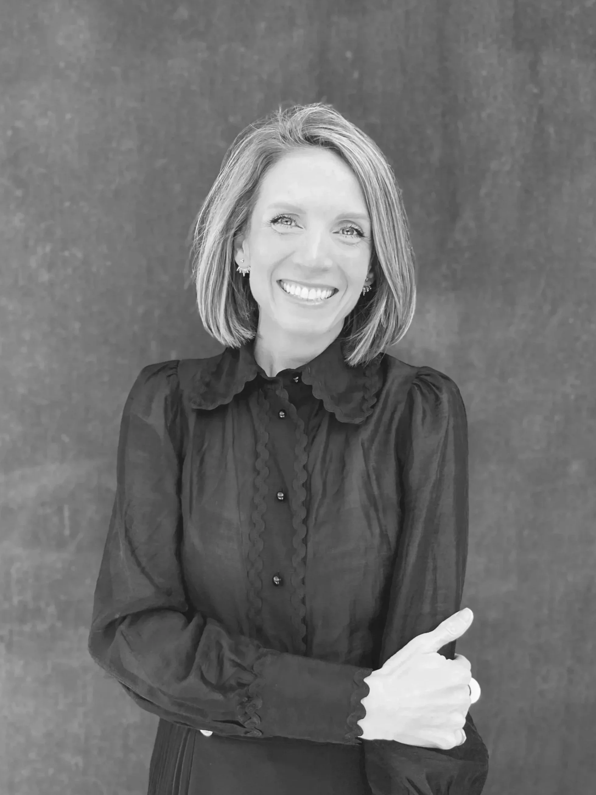 A smiling woman with shoulder-length hair wearing a dark blouse with a ruffled collar and buttons, standing against a plain background.