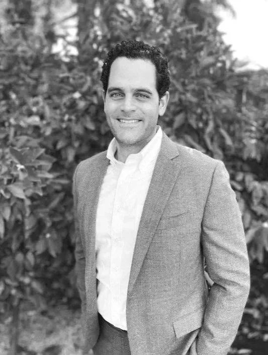 A man in a blazer and collared shirt smiling outdoors with trees in the background.
