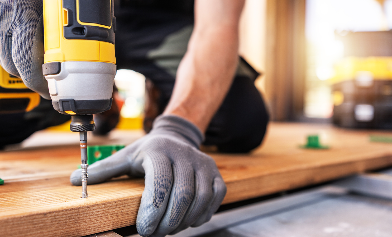 A person wearing gray gloves using a yellow and black cordless drill to screw a nail into a wooden surface, with tools and workspace in the background.