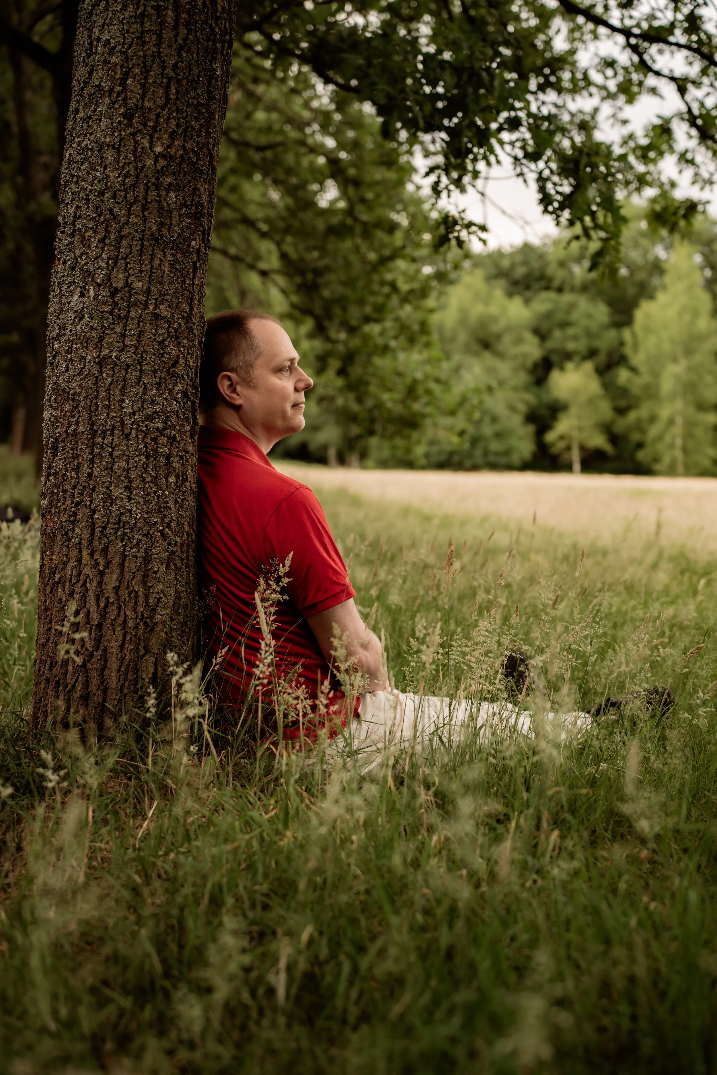 Maarten Wauters (de Nuchtere Mysticus) leunt ontspannen tegen een boom in een grasveld met bosrijke achtergrond.