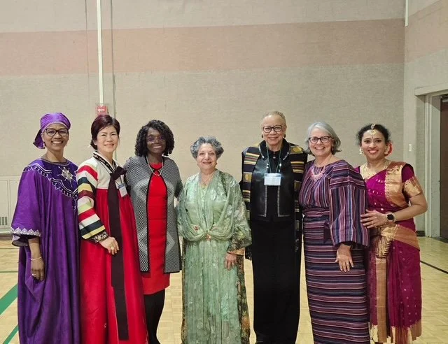 Seven women standing together indoors, dressed in colorful traditional and modern clothing, smiling at the camera.