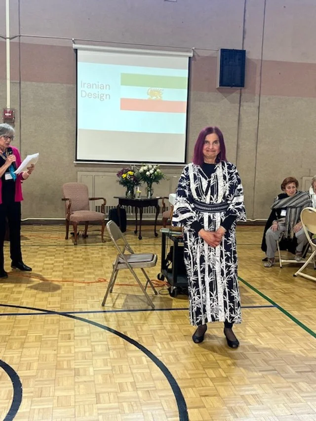 A woman with purple hair standing in a gymnasium in front of a projected slide that reads 'Iranian Design' with the Iranian flag. Several chairs and a table with flowers are behind her, and other people are seated nearby.