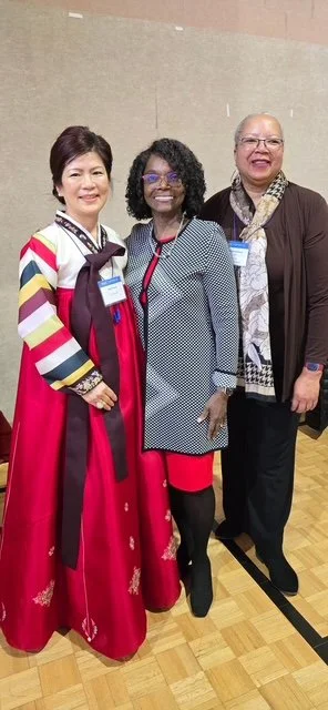 Three women standing together indoors, smiling for the photo. The woman on the left is wearing a colorful dress with a maroon overlay, the woman in the middle is wearing glasses and a gray and black patterned blazer with a red dress, and the woman on