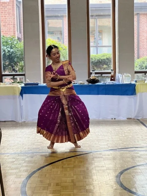 A woman in traditional Indian attire performing a dance indoors with a basketball court floor, behind her is a table with food and drinks, and large windows showing a view outside.