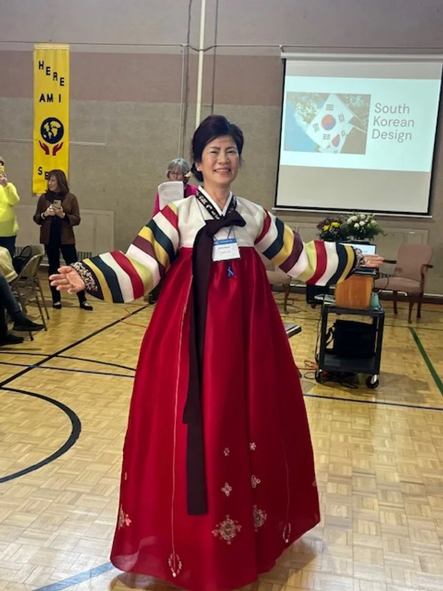 Woman wearing a traditional Korean dress, called a Hanbok, standing with arms outstretched inside a gymnasium during a cultural event.