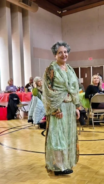 An elderly woman standing in a hall among seated women, wearing a long, colorful dress with a green scarf and smiling at the camera.