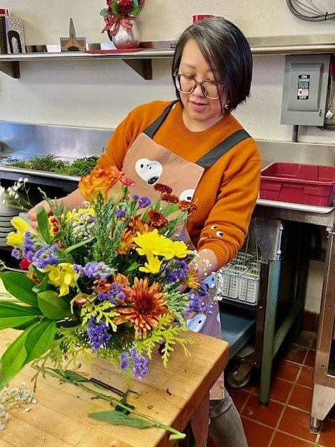 A woman with glasses and short black hair arranging a colorful flower bouquet in a kitchen.