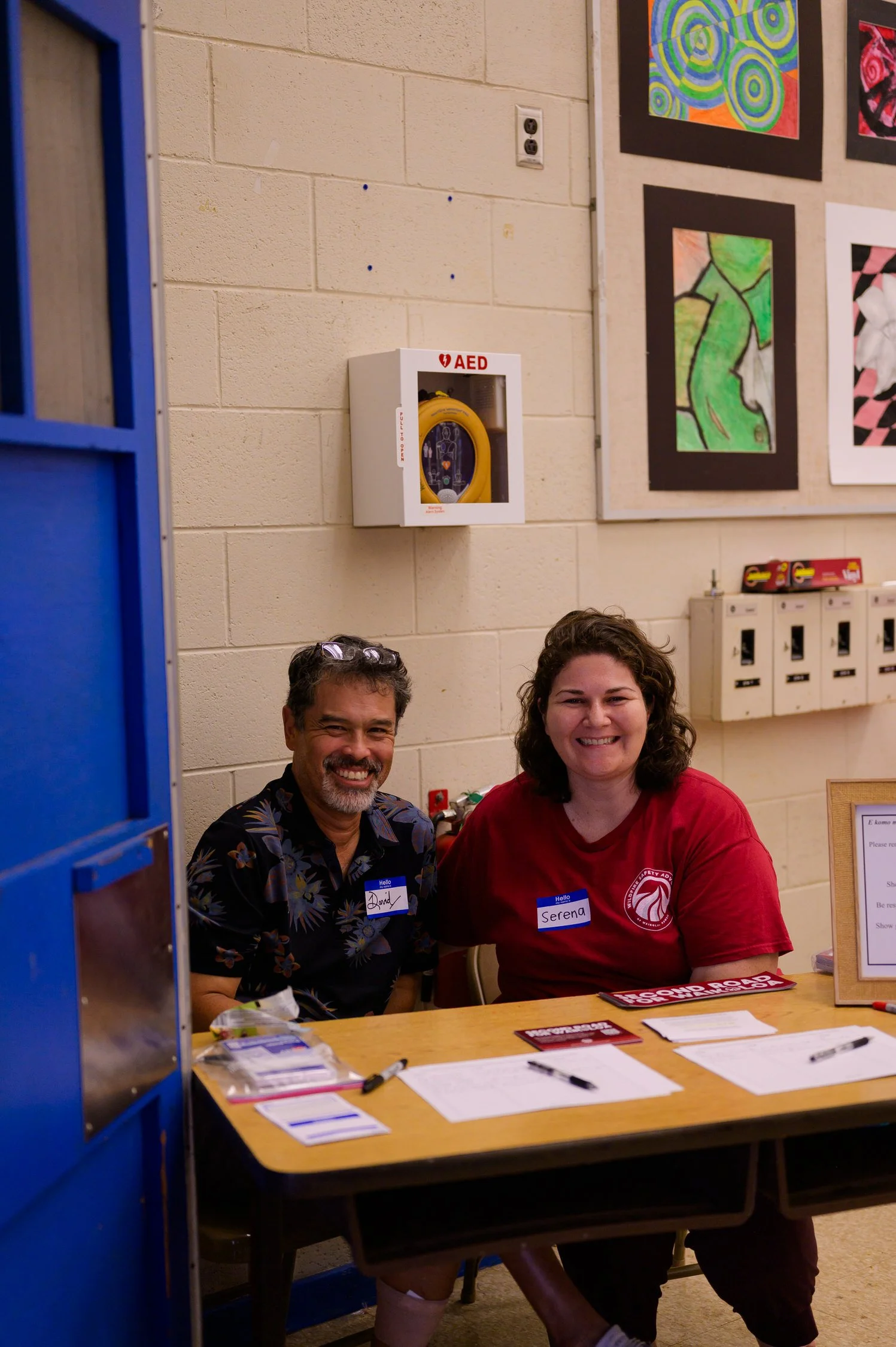 Two smiling people, a man and a woman, sitting at a table with registration forms and pens, inside a community center or school. The man has a name tag that says 'Lindy' and the woman has a name tag that says 'Serena'. Behind them on the wall are colorful paintings and an AED emergency device.
