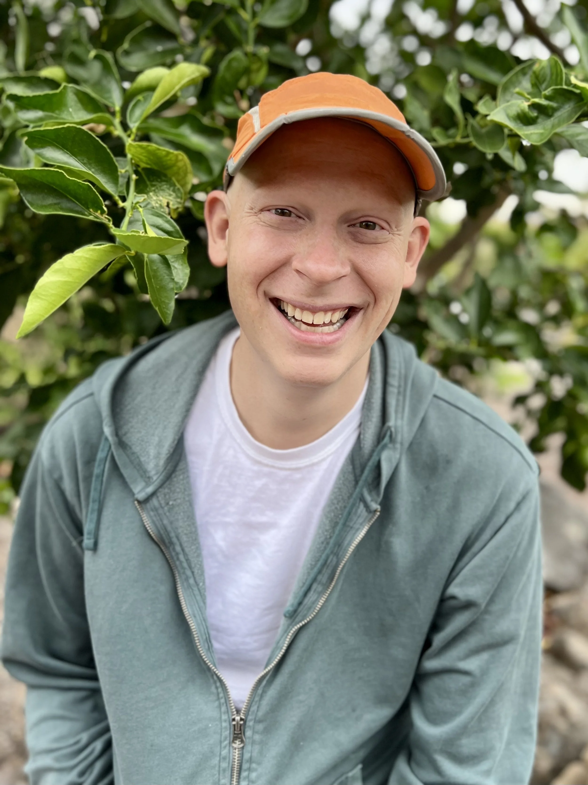 A young man smiling outdoors, wearing a beige and orange baseball cap, a white t-shirt, and a teal zip-up hoodie, standing among green leafy plants.
