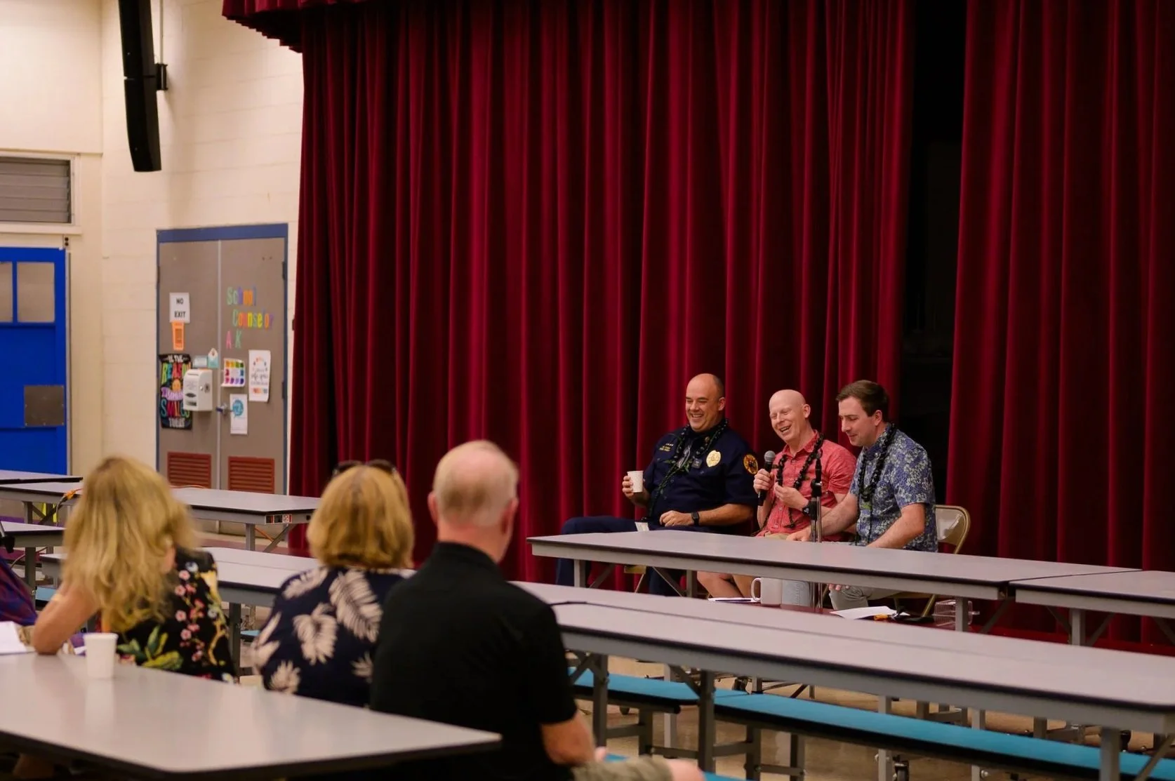 Panel of three men sitting at a table on a stage with red curtains, engaging in a discussion with an audience seated at tables.