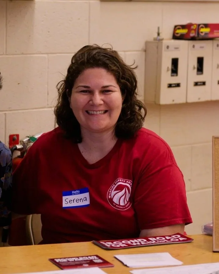 Woman with curly brown hair smiling, wearing a red shirt with a logo and a blue name tag that says 'Serena,' sitting at a table in a room with beige brick walls and postal boxes in the background.