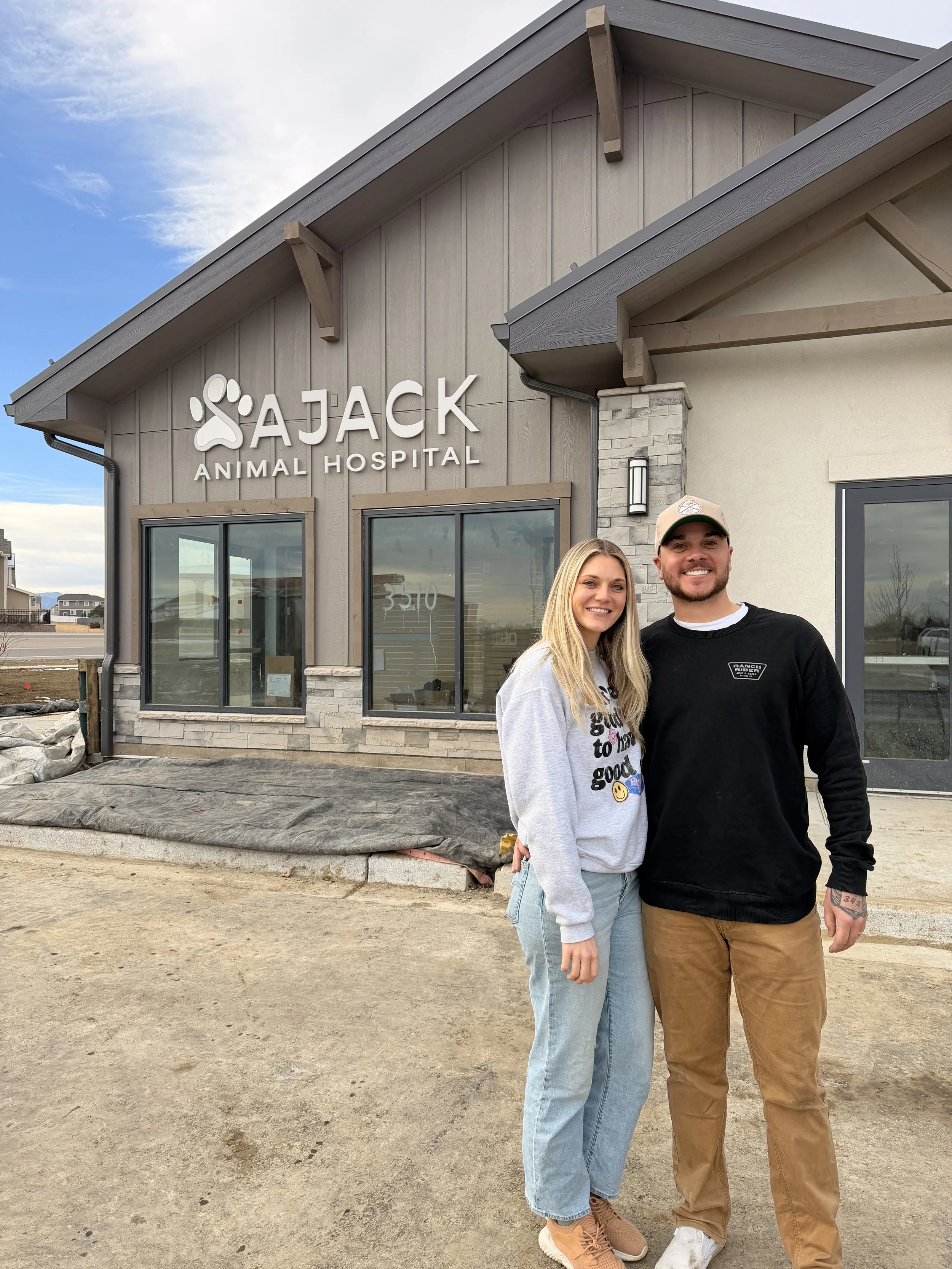 A smiling young woman and man standing in front of the Jack Animal Hospital building, which has large windows, a stone and wood exterior, and a sign with a paw print. The sky is partly cloudy.