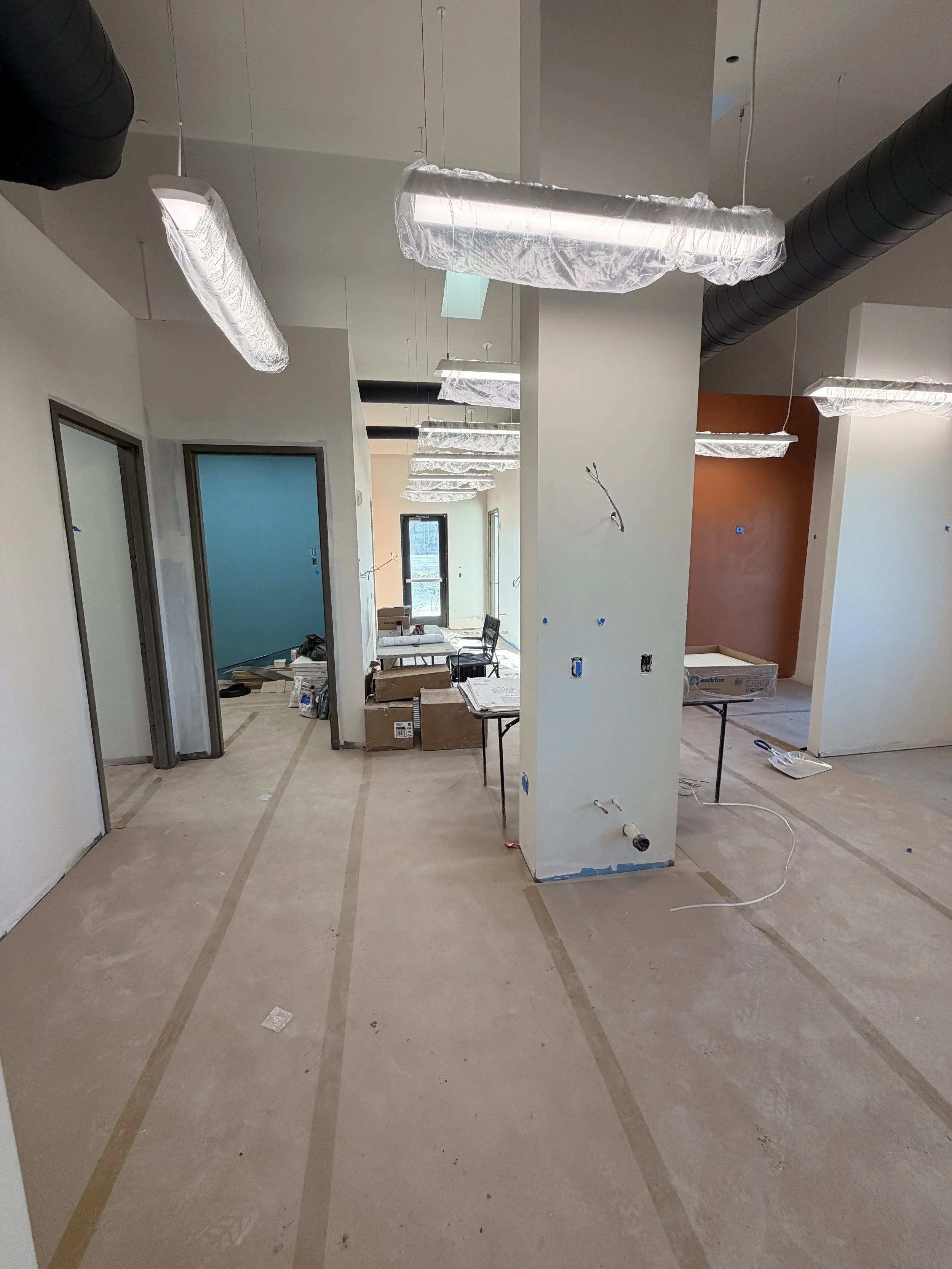 Interior of a building under construction with unfinished floors, walls, and ceiling, some walls painted in different colors, and construction materials and tools scattered around.