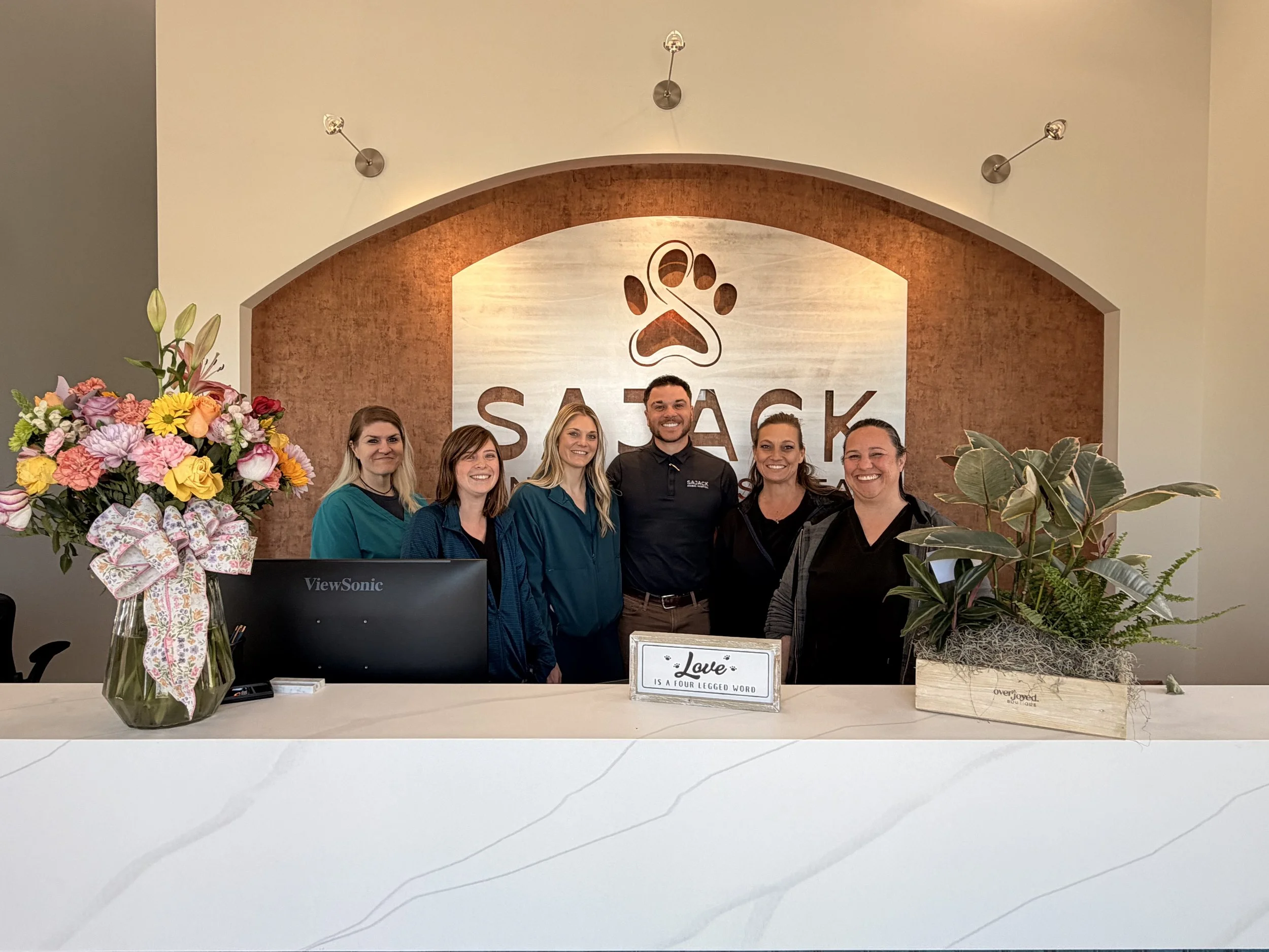 Group of six people standing behind a reception desk, smiling, with flowers on the left and a plant on the right, in front of a large logo that says "SASAK" featuring a paw print.