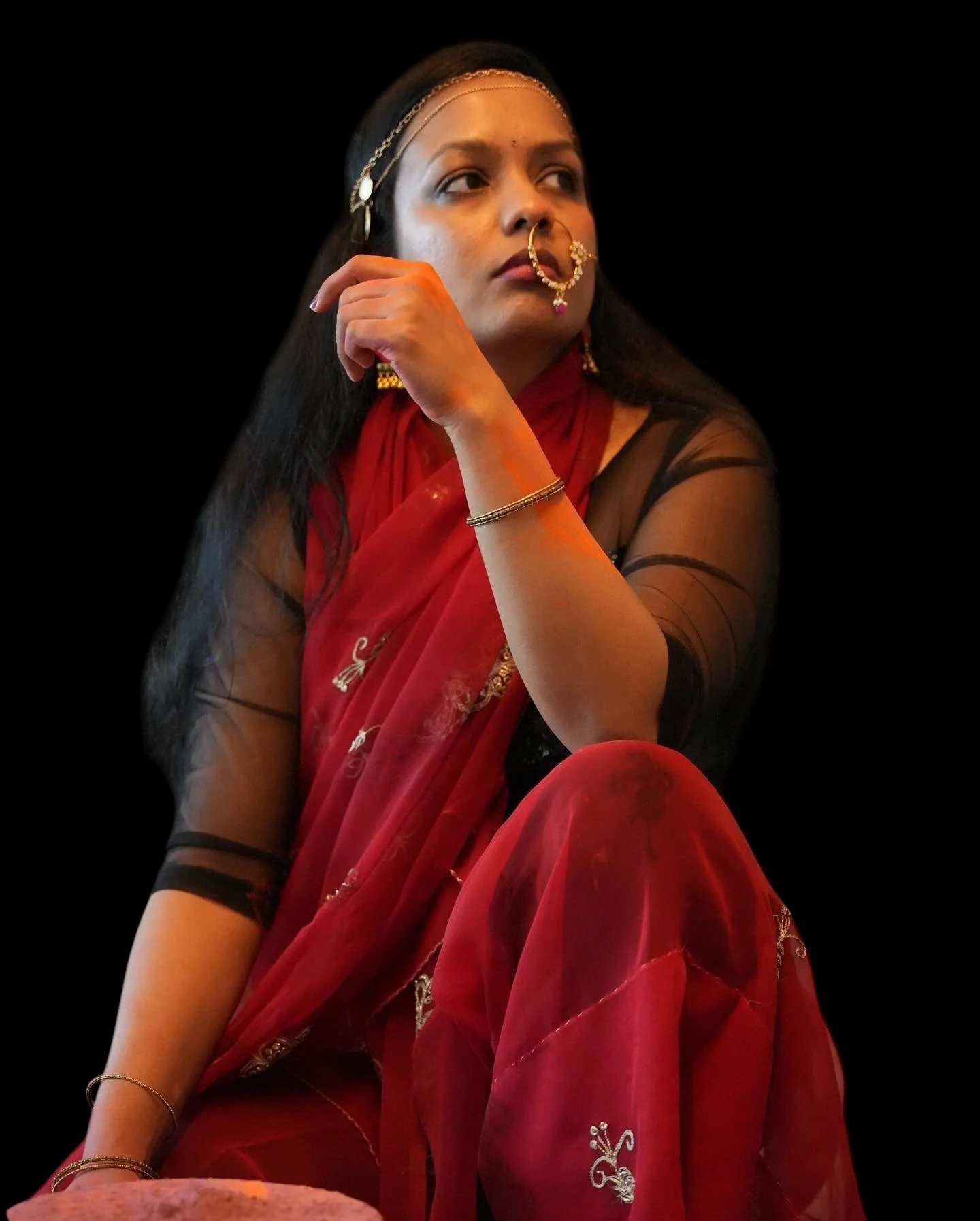 A woman dressed in traditional Indian attire, wearing a red sari with embroidery, black blouse, and gold jewelry, seated with her chin resting on her hand against a black background.