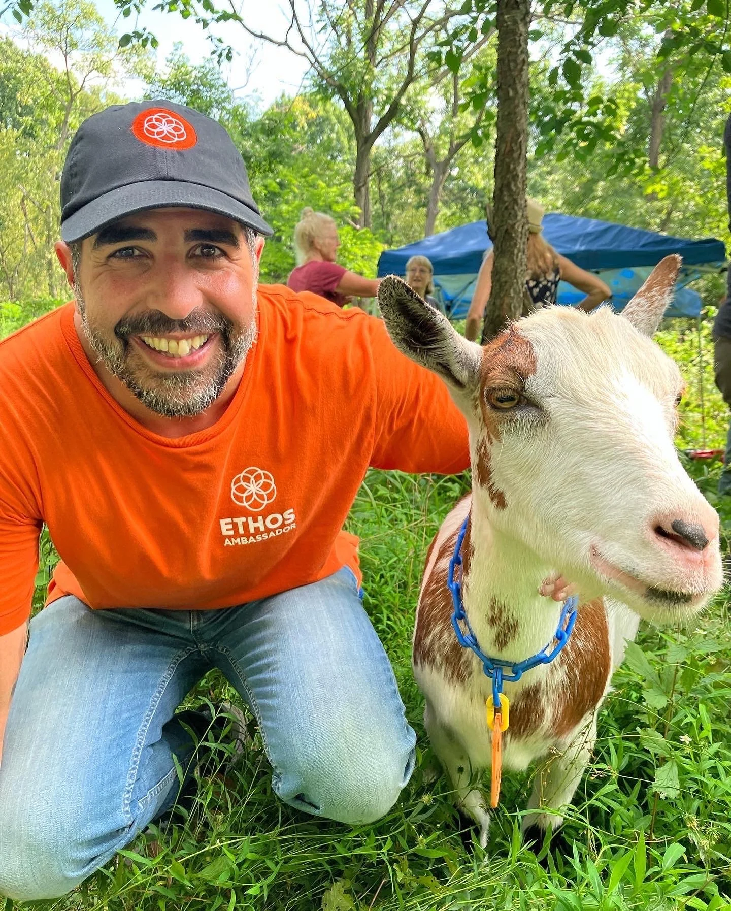 A Pittsburgher wearing an orange ETHOS Ambassador T-shirt and a gray cap is taking a selfie with a young goat in a grassy outdoor area. The goat has white and brown fur with speckled markings, blue collar, and is looking to the side.
