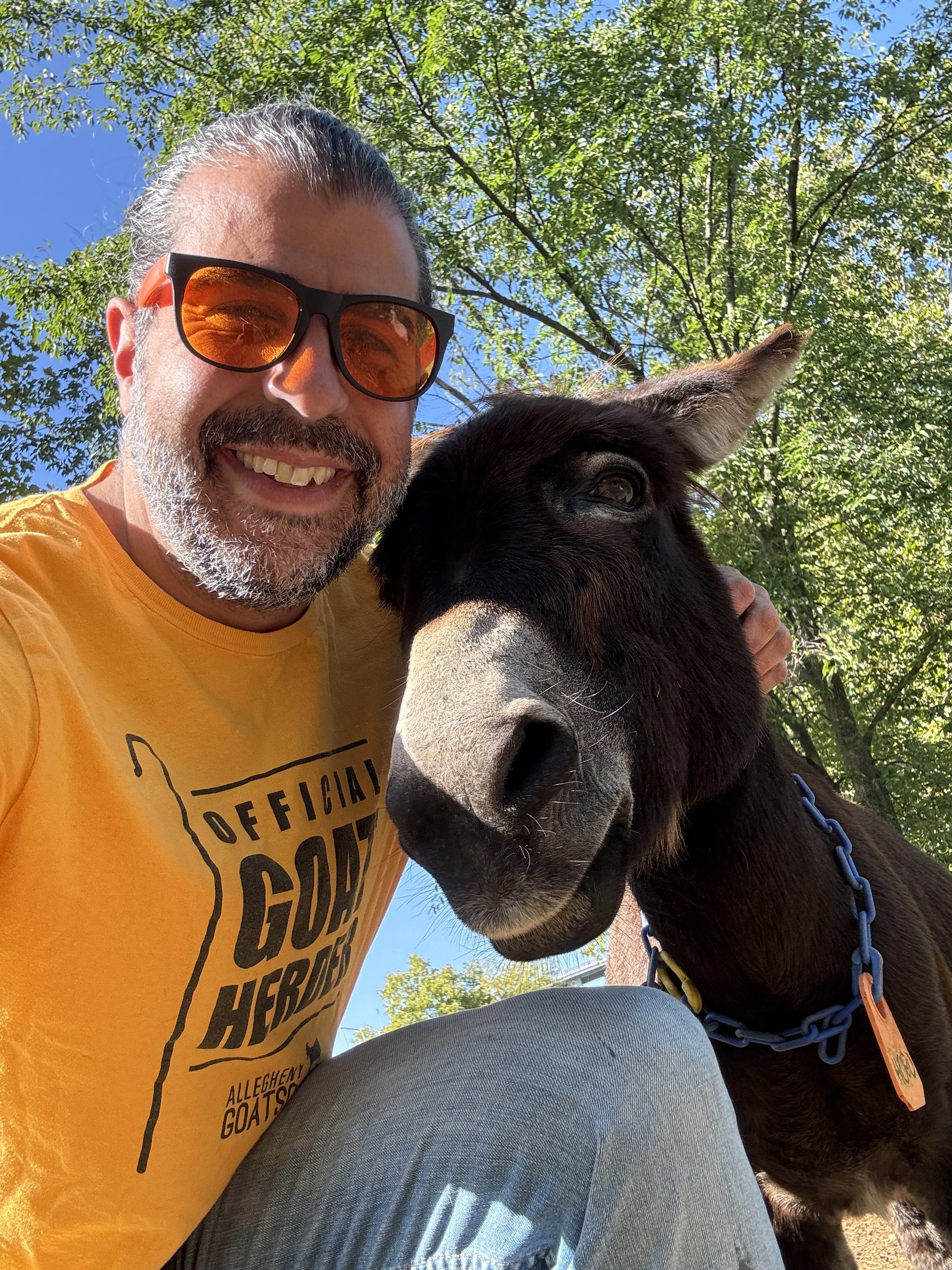 A smiling volunteer wearing sunglasses and a yellow shirt taking a selfie with a black dog outdoors on a sunny day, with green trees and blue sky in the background.