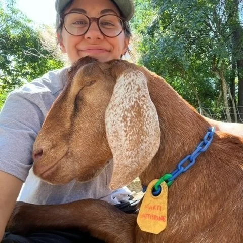 Erin wearing glasses smiling and hugging a brown goat with long, floppy ears and a blue collar with a yellow tag outdoors.