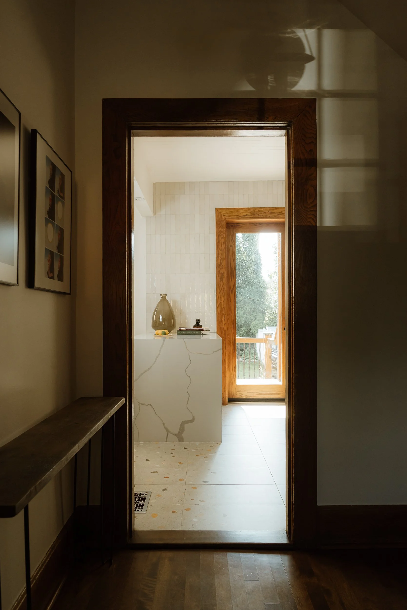 View through a doorway into a sunlit room with a marble pedestal, a vase, books, and a door leading to a balcony with trees outside.