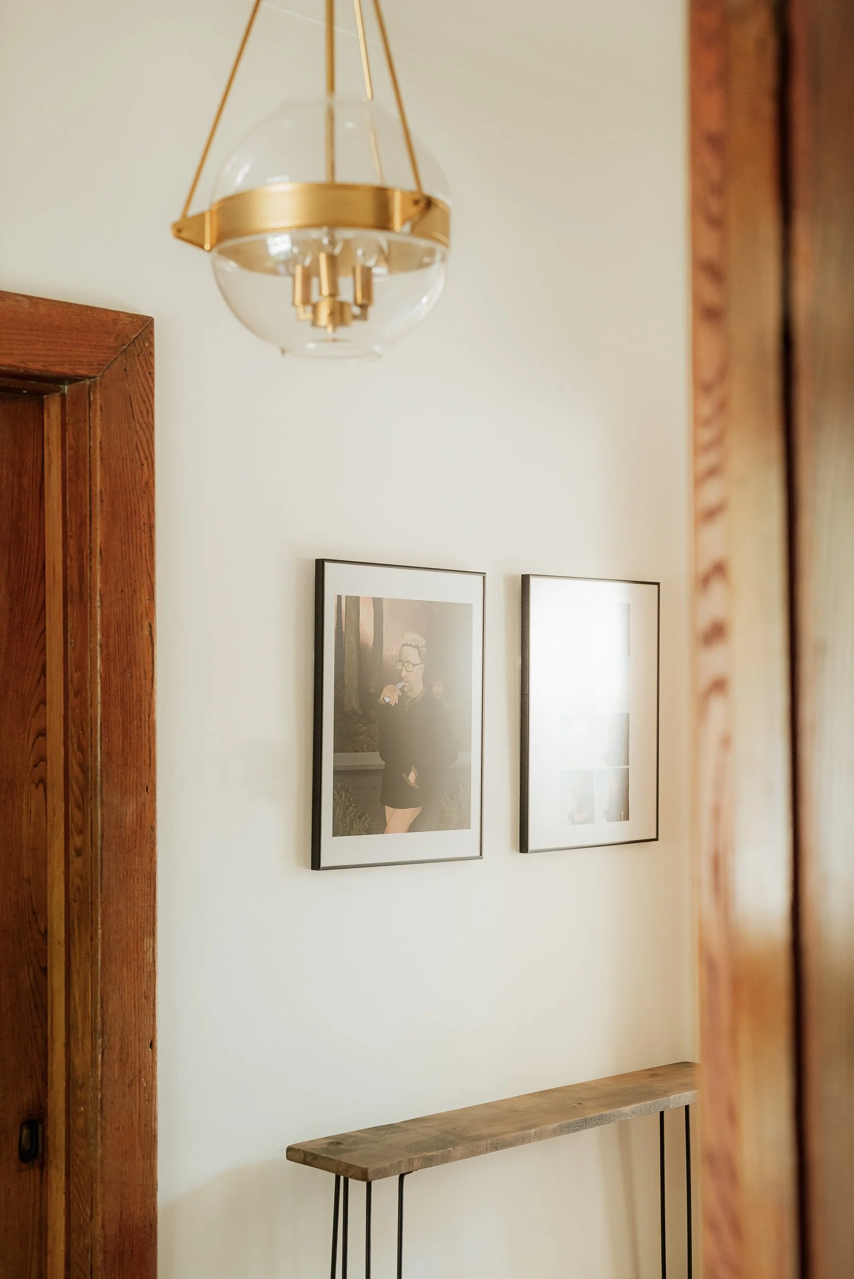 Interior view of a wall with two framed photographs, a wooden door, and a wooden table beneath the photos, with a modern hanging light fixture above.