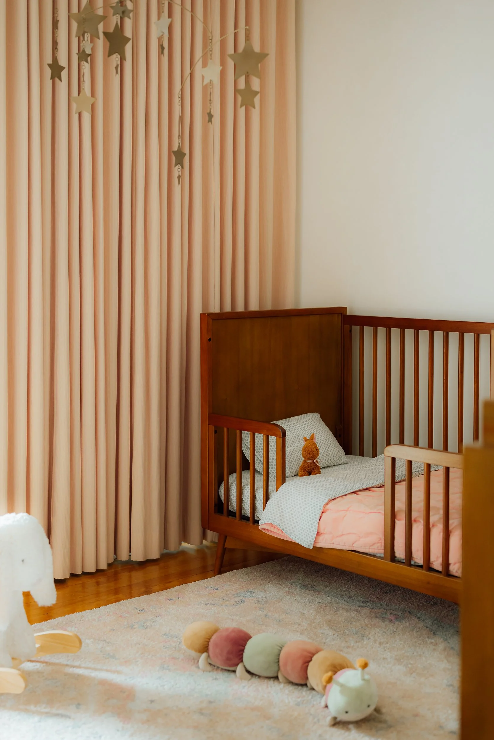 Nursery room with wooden crib, stuffed animal toy, pink and white bedding, plush caterpillar on rug, beige carpeting, pink curtains, and star decorations hanging from ceiling.