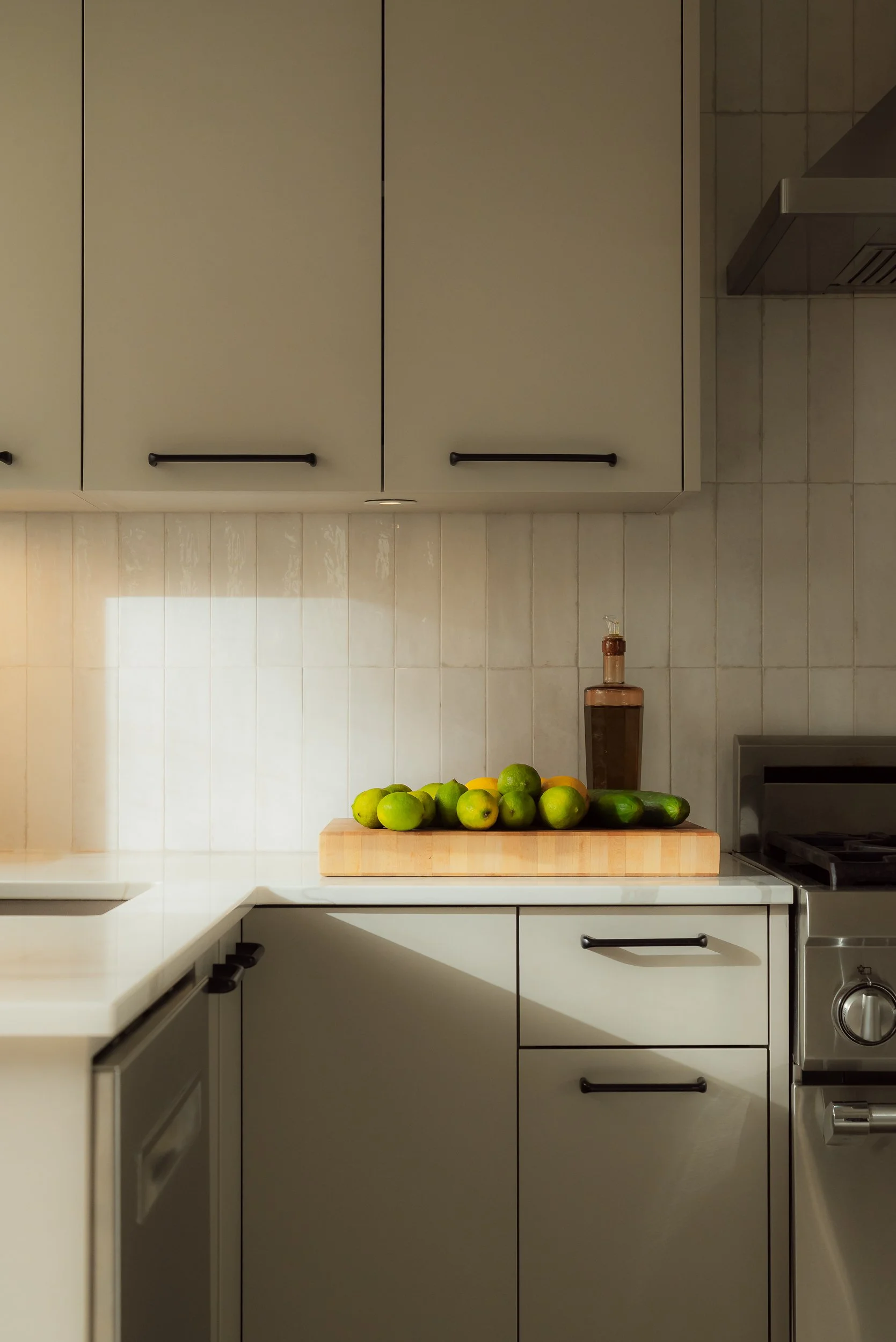 Kitchen with white cabinets, a white countertop, a cutting board with limes and cucumbers, a brown bottle, and a stove.