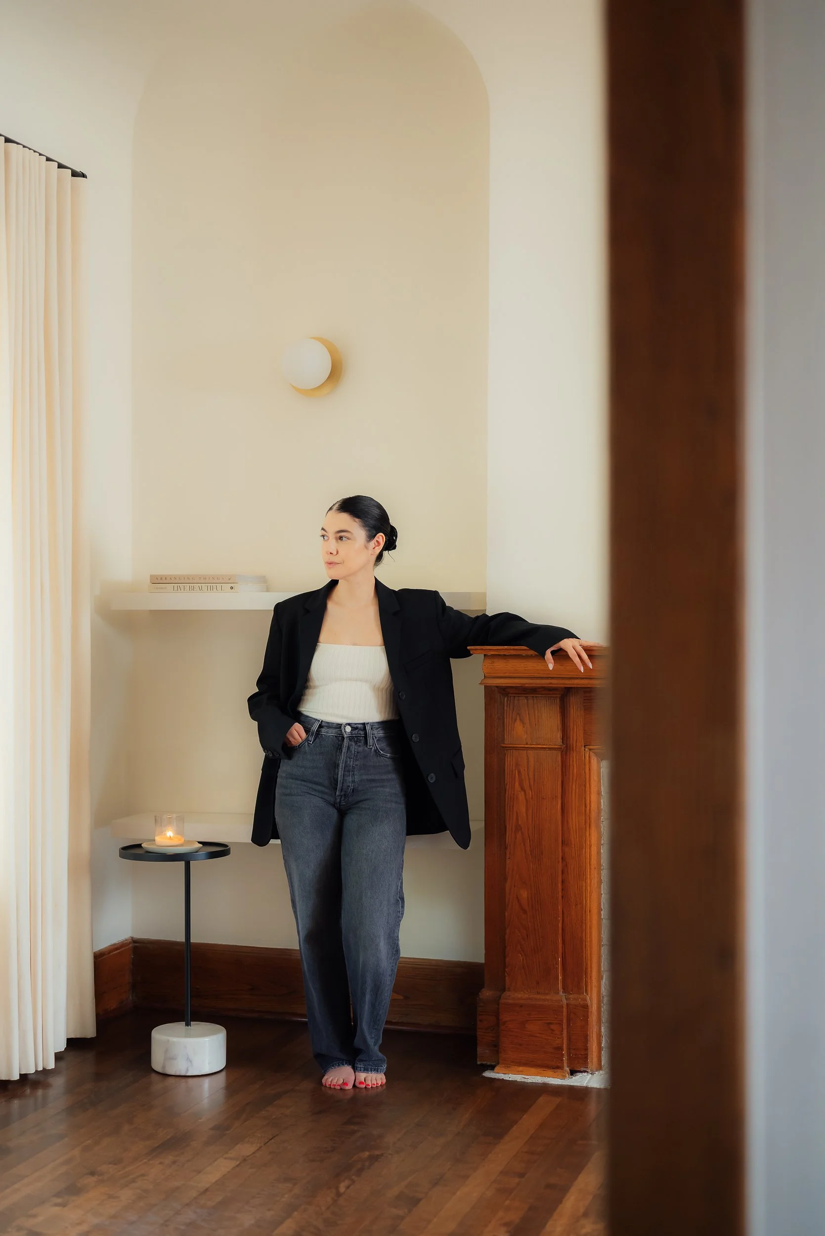 A woman standing in a room, leaning against a wooden column, wearing a black blazer, white top, and jeans, with dark hair pulled back, near a side table with a candle, in a minimalist decor setting.