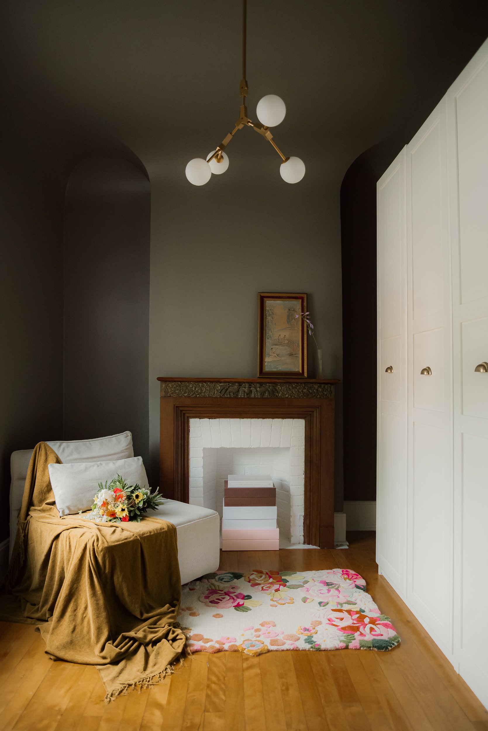 A cozy corner with a white armchair draped in a brown blanket, decorated with a bouquet of flowers on it. There is a colorful floral rug on a wooden floor, a fireplace with a wooden mantel, a framed picture above the mantel, and a white wardrobe on the right side. A modern chandelier hangs from the ceiling.